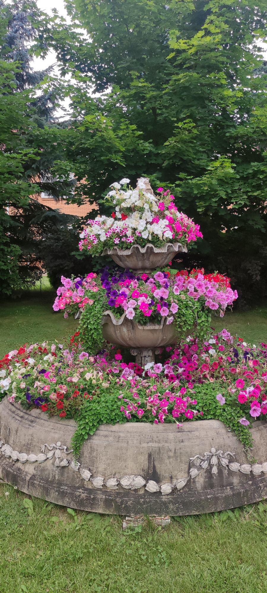 The image shows a three-tiered, weathered stone fountain overflowing with a vibrant display of petunia flowers. The fountain is situated outdoors on a grassy lawn, with lush green trees forming a dense backdrop. The flowers are a mix of colors, including white, pink, purple, and red, with some trailing greenery cascading down the sides. The stone fountain itself has decorative carvings along its base. The scene suggests a well-maintained garden or park setting. The location is identified as Rosinka, Russia, though no specific landmarks or identifying features of that city are visible within the frame. There are no people or animals present in the image.