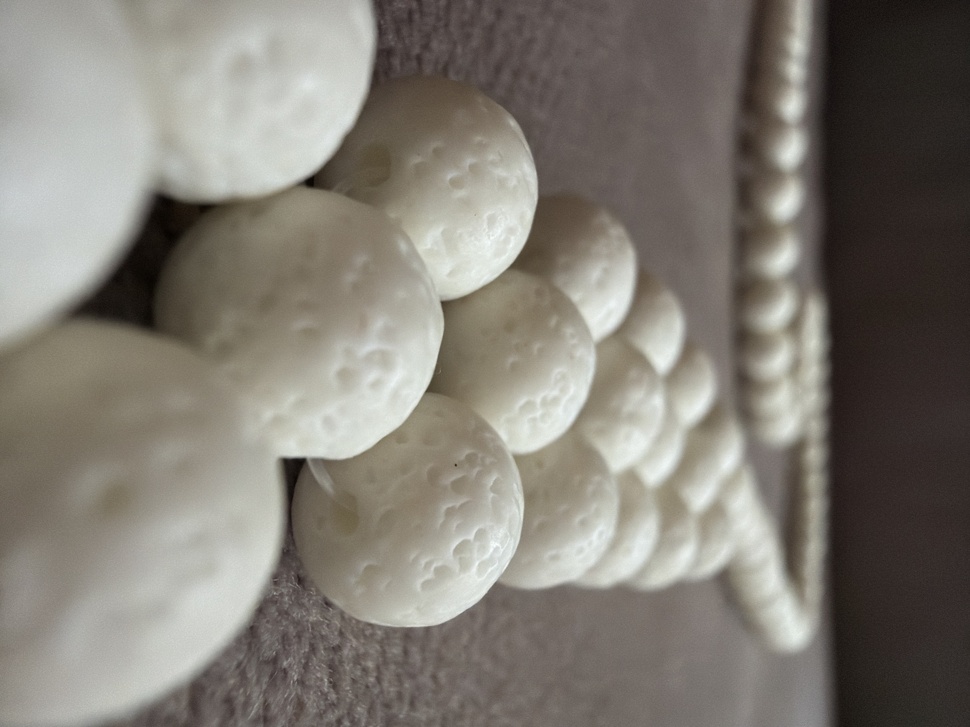 The image presents a close-up view of numerous small, white, spherical beads laid out on a textured, muted grey-brown fabric surface. The beads are arranged in a diagonal line across the frame, suggesting they are strung together, possibly as a rosary or prayer beads.
The beads in the foreground are in sharp focus, revealing a distinct, irregular, and slightly porous or textured surface, giving them an organic or crafted appearance. They are off-white or creamy in color. As the line of beads recedes into the background, they become progressively smaller and less distinct due to a shallow depth of field, with a blurred string extending along the right side of the frame. The soft lighting highlights the three-dimensional form and surface details of the individual beads. No specific activity is depicted, nor are there any people or visible text. The setting appears to be indoors.