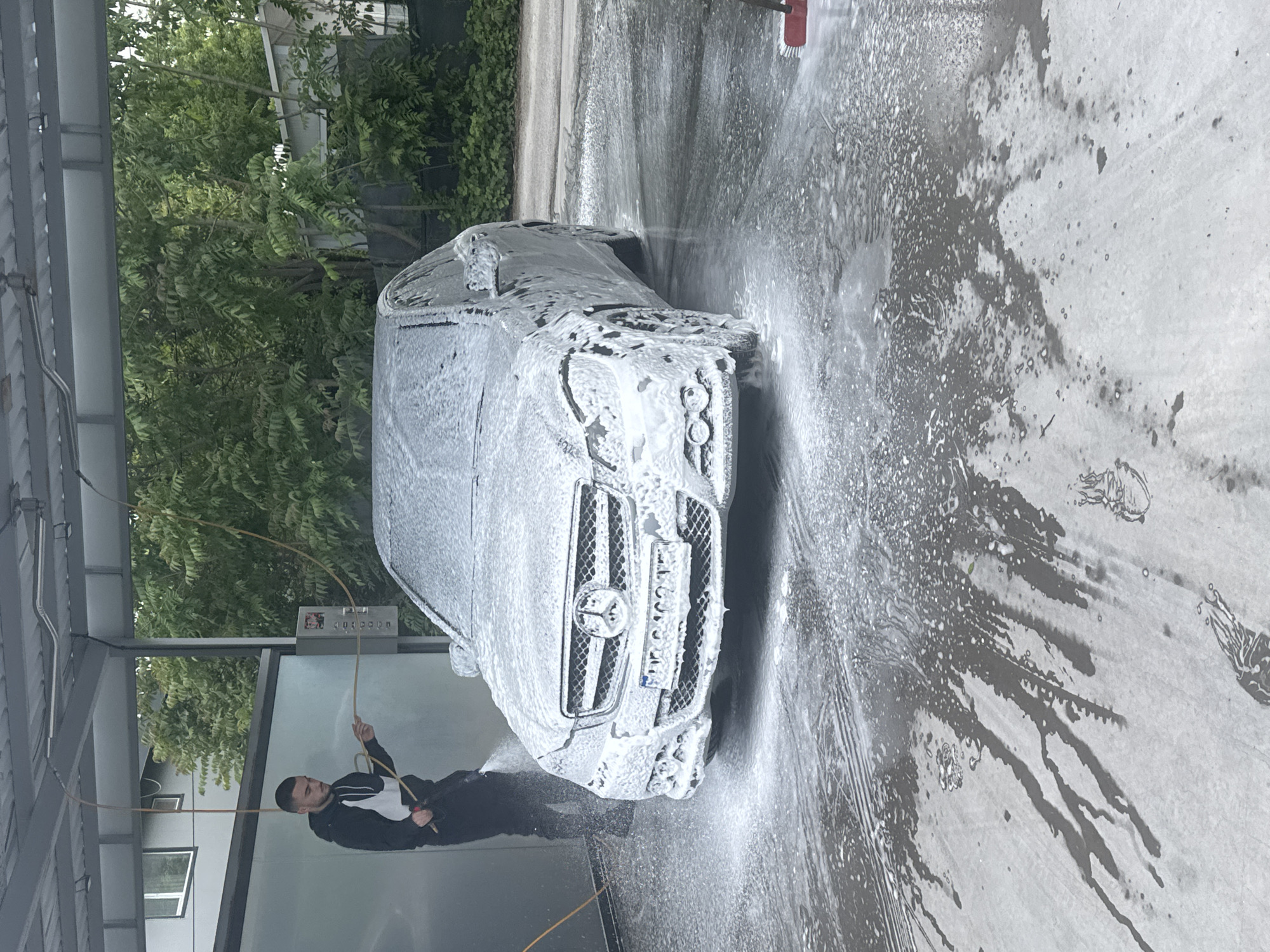 A man is washing a car at a car wash. The car is covered in soap suds. The man is holding a hose and spraying water on the car. The car wash is located in a city, likely Plovdiv, Bulgaria. Trees and parts of buildings are visible in the background.