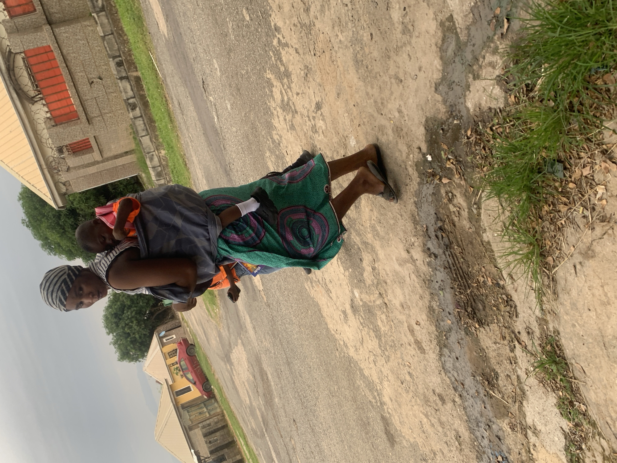 A woman carries a child on her back in a grey baby sling while walking on a dirt and asphalt road. She wears a striped headscarf, a striped shirt, and a green patterned skirt. The child is wearing an orange shirt and white socks. Behind them, there are two buildings with yellow roofs and a red car parked in front of one. The scene appears to be in a residential area in Abuja, Nigeria. The sky is overcast.