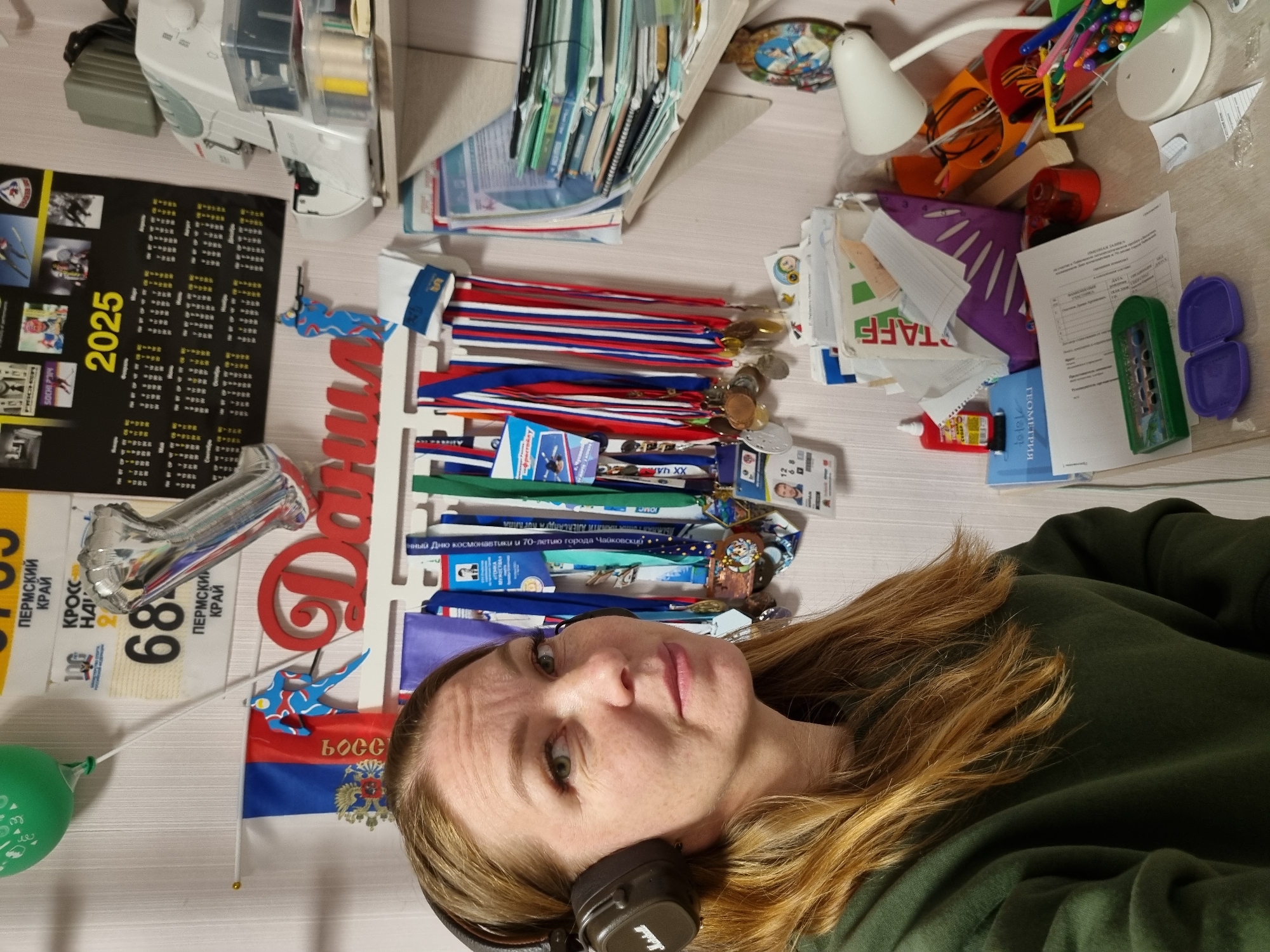 The image captures a woman with long reddish-brown hair and a neutral expression, wearing a dark green long-sleeved top and black Marshall over-ear headphones. She is positioned in the foreground, with her head slightly tilted.
The background reveals a cluttered indoor space, likely a desk or table. A central feature is a rack displaying numerous sports medals with red, white, and blue ribbons, signifying athletic achievements. A small Russian flag with the double-headed eagle emblem is visible, alongside a red wooden cutout spelling "Даша" (Dasha).
Other objects on the surface include a white sewing machine, a white desk lamp, a black calendar for "2025," and a silver foil balloon partially showing "68" next to text "Пермский Край" (Perm Krai). A green balloon is also partially visible. Stationery items such as pen holders filled with pens and markers, a red pencil sharpener, and various papers are scattered across the table. Among the papers, one is labeled "STAFF" and another, "ЗАЧЁТНАЯ ЗАЯВКА" (Entry Application). A blue book titled "ГЕОМЕТРИЯ" (Geometry) is also present.
Text on some medal ribbons indicates specific events or locations, including "70-летию города Чайковский" (70th anniversary of Chaikovsky) and "Дню космонавтики" (Cosmonautics Day), firmly placing the context in Russia, specifically Chaikovsky. The scene suggests a personal workspace, reflecting hobbies, accomplishments, and daily activities.