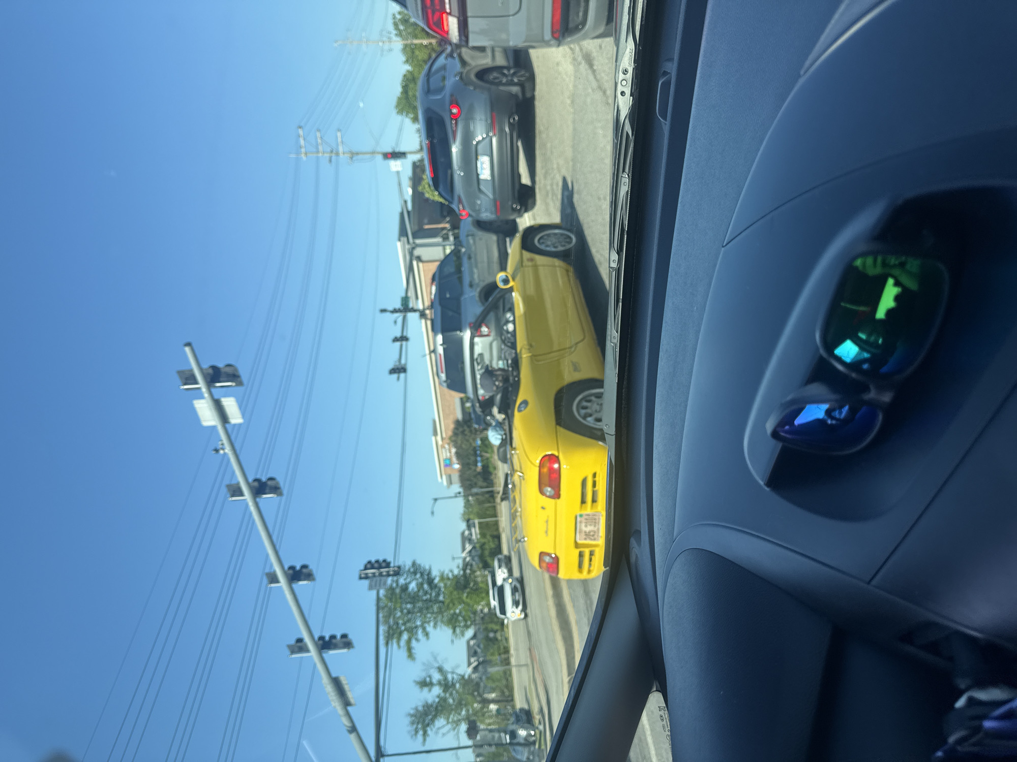 The image, taken from inside a vehicle in Morton Grove, Illinois, captures a street scene with several cars stopped at a traffic light. A bright yellow convertible is prominently visible in the adjacent lane, with its license plate reading "216 3XM." Other vehicles, including a grey SUV, are lined up behind it. Traffic lights and power lines extend vertically along the left side of the frame, contrasting with the clear blue sky. The interior of the vehicle from which the picture was taken is visible, with the dashboard and a pair of sunglasses in view. There are no identifiable people within the frame, only the objects and environment indicate the scene.