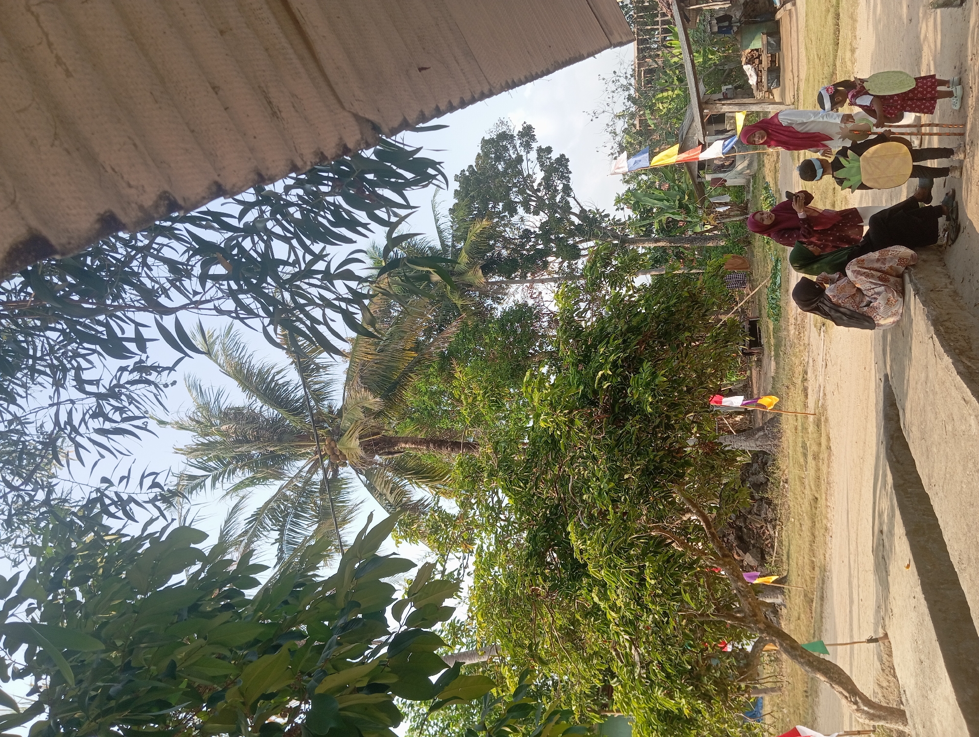 The image captures a rural scene in Dente Teladas, Indonesia, under a clear sky with sunlight illuminating the area. The foreground features a concrete structure, possibly a raised walkway or seating area, with two children and three adults gathered. One child, dressed in a costume resembling a pineapple, stands facing the adults. Another child, in a patterned dress, holds a prop. The adults are dressed in traditional clothing, with two of them wearing hijabs. One adult is looking at a smartphone. Behind them, a dirt path winds through a landscape dotted with lush green trees, including a prominent palm tree. Small flags in red, white, yellow, and purple are visible, planted in the ground. In the background, a rustic wooden structure with a corrugated roof is present, along with piles of wood and what appear to be simple dwellings. The overall atmosphere suggests a casual outdoor gathering in a tropical, rural setting.