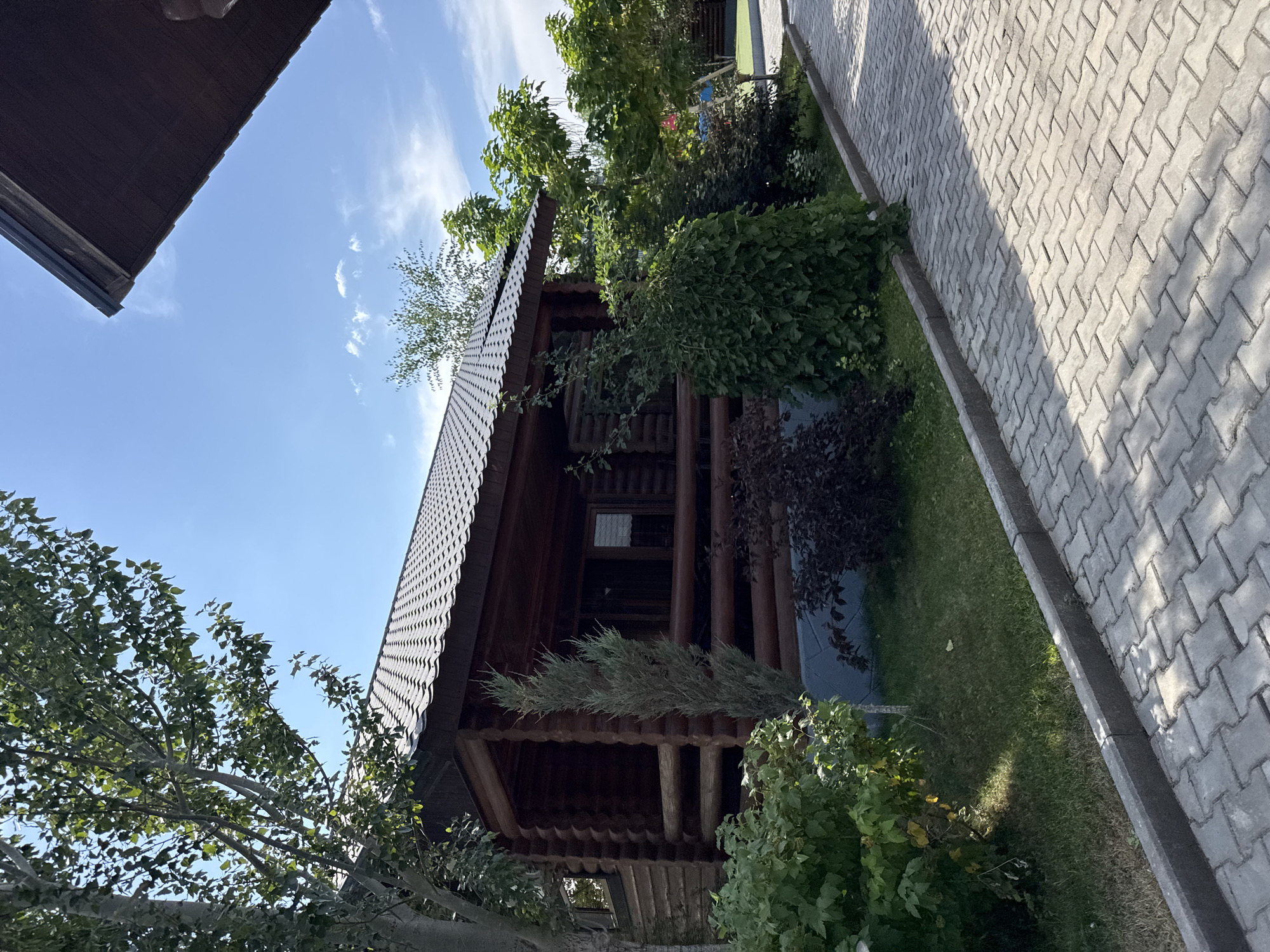 The image depicts a log cabin with a dark, textured roof, situated in a green, landscaped area. A paved walkway, made of interlocking gray blocks, runs along the side of the cabin. Lush green bushes and trees surround the structure, with some casting shadows on the walkway. The sky above is a clear blue with a few scattered white clouds. The scene appears to be a serene outdoor setting, likely a recreational area or a dwelling in a natural environment. It is not possible to determine the specific location in the city of Ақши, Kazakhstan from the visual information provided.