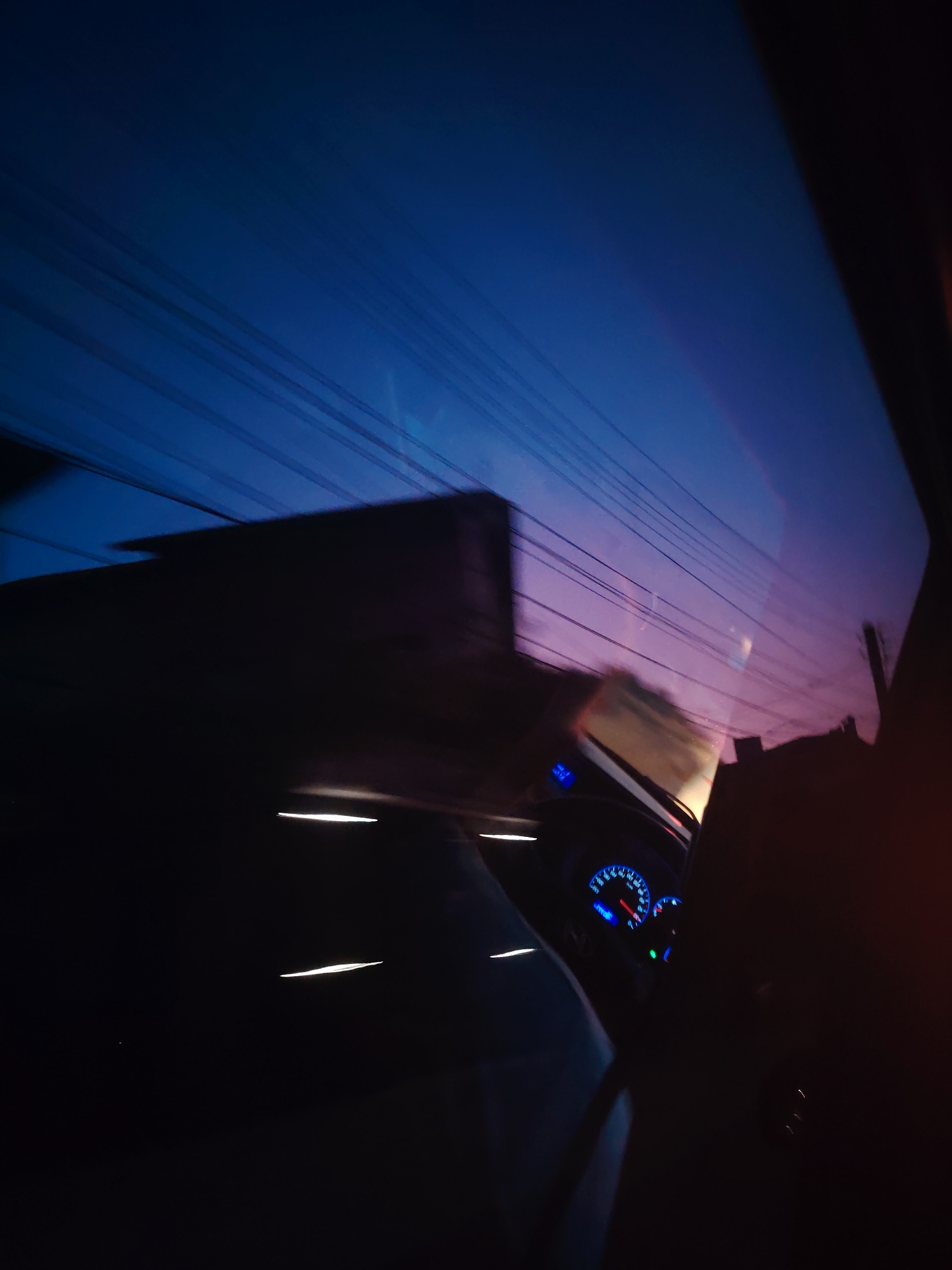 This is a low-light, motion-blurred image taken from inside a vehicle. The foreground is dominated by the dark interior of the car, with some illuminated elements such as dashboard lights and streaks of light from the car's movement. The dashboard features illuminated gauges, with blue and white lights visible. The windows of the car reflect the external environment, which appears to be a twilight or early night sky with a gradient of deep blue to purple. Several parallel lines, likely power lines or overhead wires, are visible stretching across the sky. A large, dark, and blurred object, possibly a building or another vehicle, is visible in the mid-ground, also appearing to be in motion. The overall impression is that of a car traveling at night or in very low light conditions, with the surrounding environment blurred by motion.