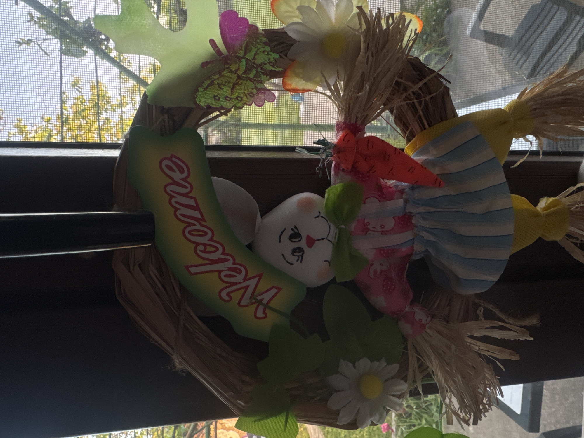 The image shows a decorative wreath hanging in front of a window with a screen. The wreath is made of straw and decorated with several elements: a white bunny head with a face, a green "Welcome" sign, a butterfly, two straw-like bundles tied with yellow fabric, a straw-like bundle tied with pink fabric and a carrot, white flowers with yellow centers, and green leaves. The window screen and a view of trees with yellowing leaves outside are visible in the background. There is no discernible information in the image to confirm that the location is in the city of Lehchevo, Bulgaria. There are no people visible in the image.