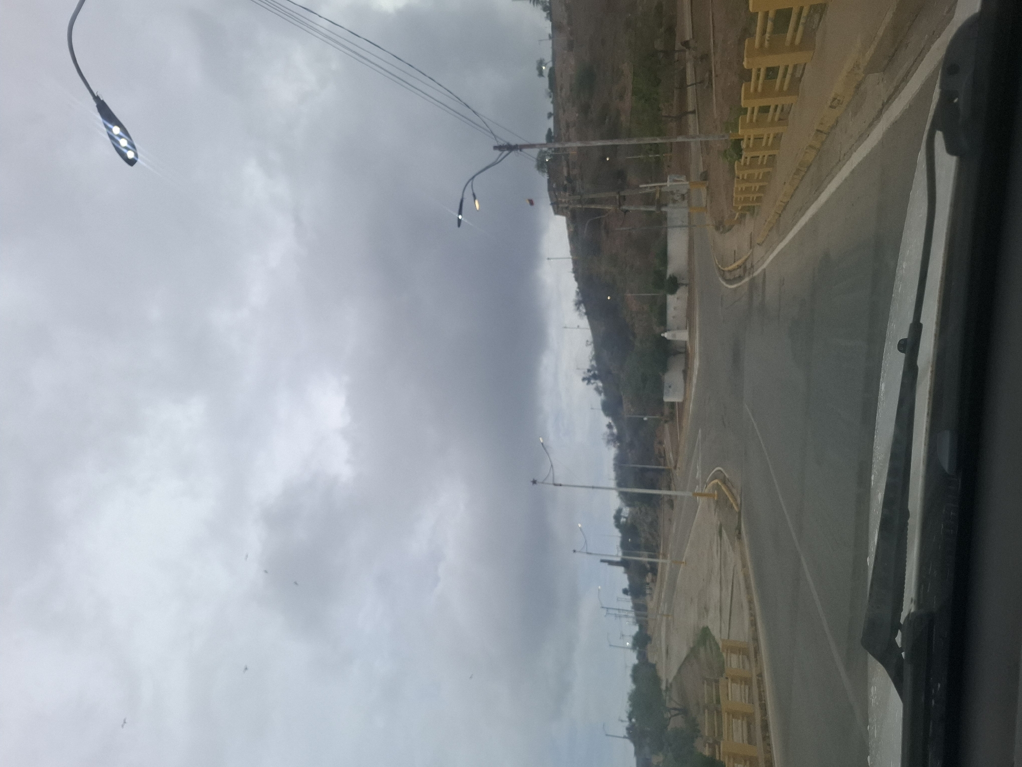 The image shows a road with streetlights in Juan Griego, Venezuela. The road curves to the right, bordered by a yellow barrier on the left and a hillside on the right. The sky is overcast and appears to be a cloudy day, possibly late afternoon. Several birds are visible in the sky, flying amongst the clouds. The scene suggests a somewhat desolate or undeveloped area, with vegetation visible on the hillside and near the road. The perspective seems to be from a vehicle moving along the road.
