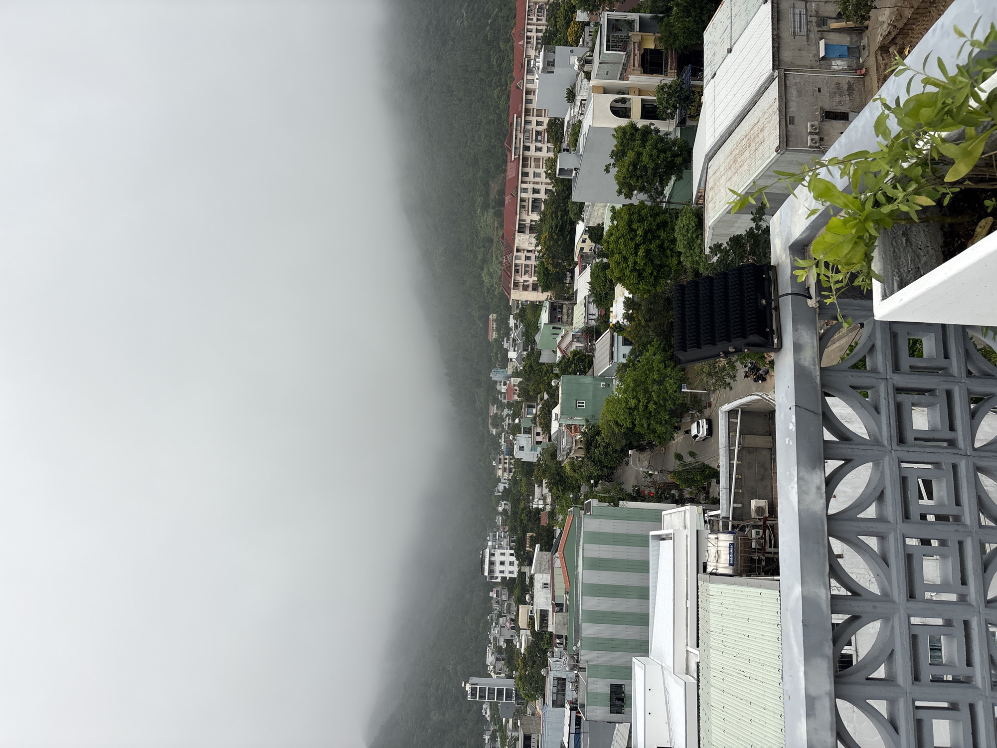 This high-angle, landscape-oriented image captures a city view in Đà Nẵng, Vietnam, under an overcast and foggy sky.
The scene is divided into a misty natural landscape on the left and a dense urban environment on the right. On the far left, a large, tree-covered mountain range is heavily obscured by thick fog or mist, blending into the uniformly light grey sky.
The midground and center of the image reveal a vibrant city with numerous residential and commercial buildings of varying heights and architectural styles. Many structures feature light-colored facades, and roofs display a mix of red tiles, grey, and distinctive green-and-white corrugated metal. Lush green trees are interspersed throughout the urban fabric, providing a contrast to the buildings. Streets are visible between buildings, with a few cars and motorcycles parked or moving. Rooftops display utilitarian elements such as air conditioning units and stainless steel water tanks; one tank clearly shows the text "SINH THAI" and possibly "Năng lượng mặt trời" below it. A prominent large black spotlight or similar device is visible on a rooftop in the mid-right.
In the immediate foreground, to the right, a grey concrete balcony railing with an intricate geometric pattern of circles and squares is visible. A potted plant with bright green leaves grows from a planter box situated on or near the railing.
The time of day appears to be daytime, though the heavy overcast conditions and fog create a subdued, tranquil atmosphere. The pervasive mist over the mountain suggests humid weather.