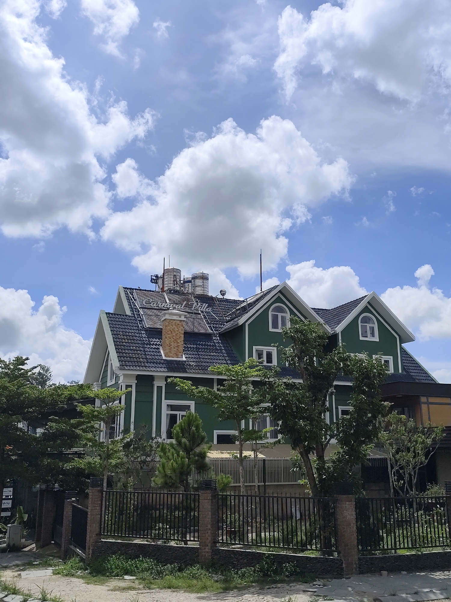 The image depicts a building called "Carnival Villa" in Da Lat, Vietnam. The villa is a two-story green structure with a dark gray roof, white trim, and arched windows on the upper floor. A chimney is visible on the roof, along with metallic tanks and piping. The building is partially obscured by green trees and a black metal fence with brick pillars. The ground in front of the fence has some grass and vegetation. The sky above is bright blue with scattered white clouds.