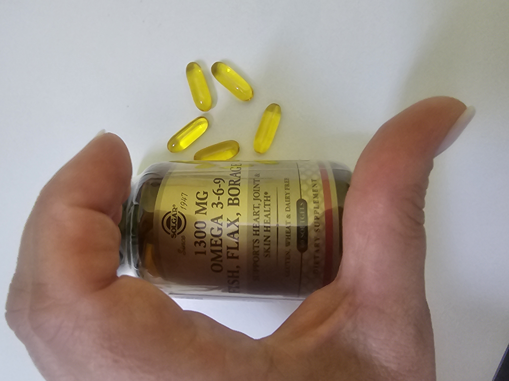 The image shows a hand holding a bottle of Solgar Omega 3-6-9 dietary supplement capsules. The bottle is amber-colored, and the label indicates it contains 1300mg of omega fatty acids derived from fish, flax, and borage. Several yellow, oblong capsules are scattered near the top of the bottle. The background is a plain, light surface. The location is not possible to determine from the image.