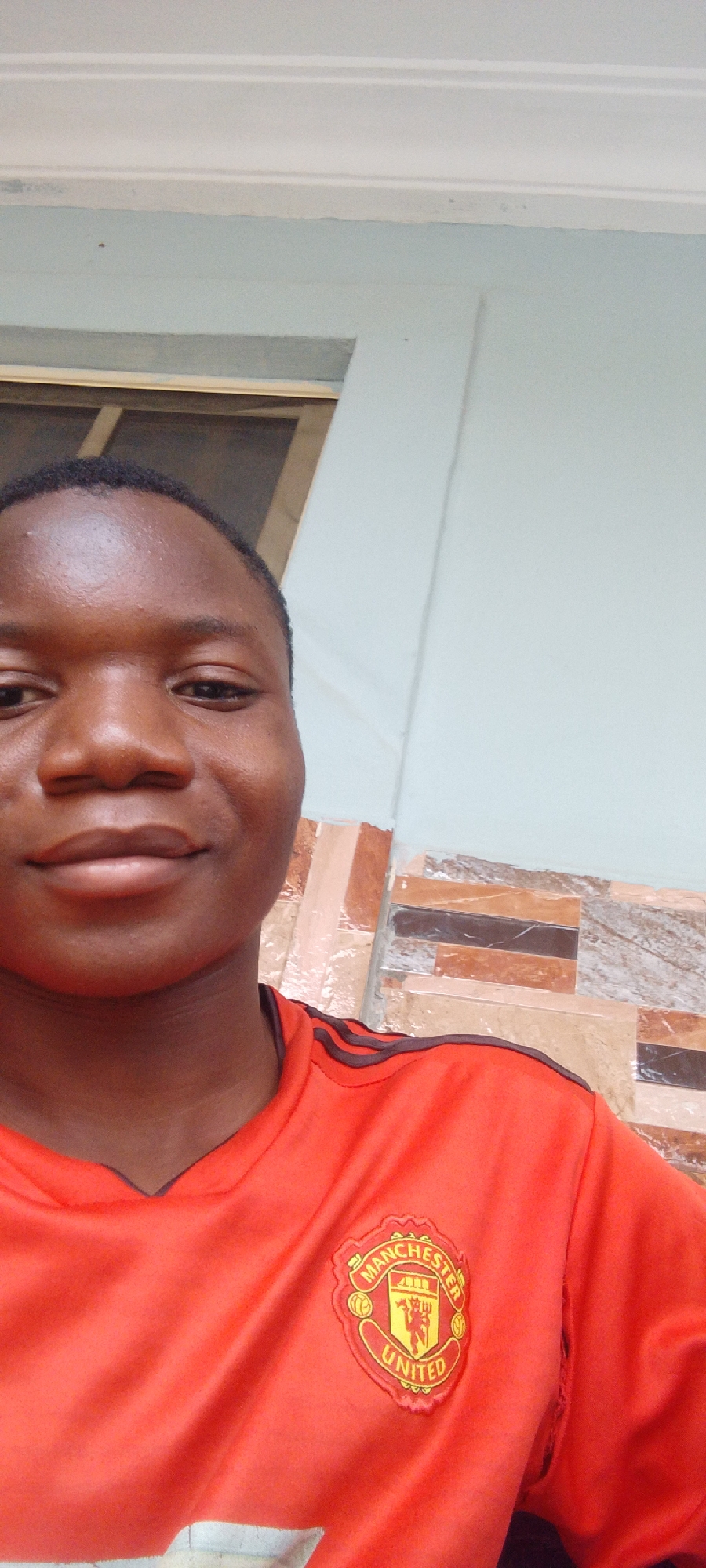 A person wearing a red Manchester United soccer jersey is facing the camera. The person is likely indoors. The background consists of a light-colored wall and a section with brown and tan tiles. The location is Unknown, Unknown.