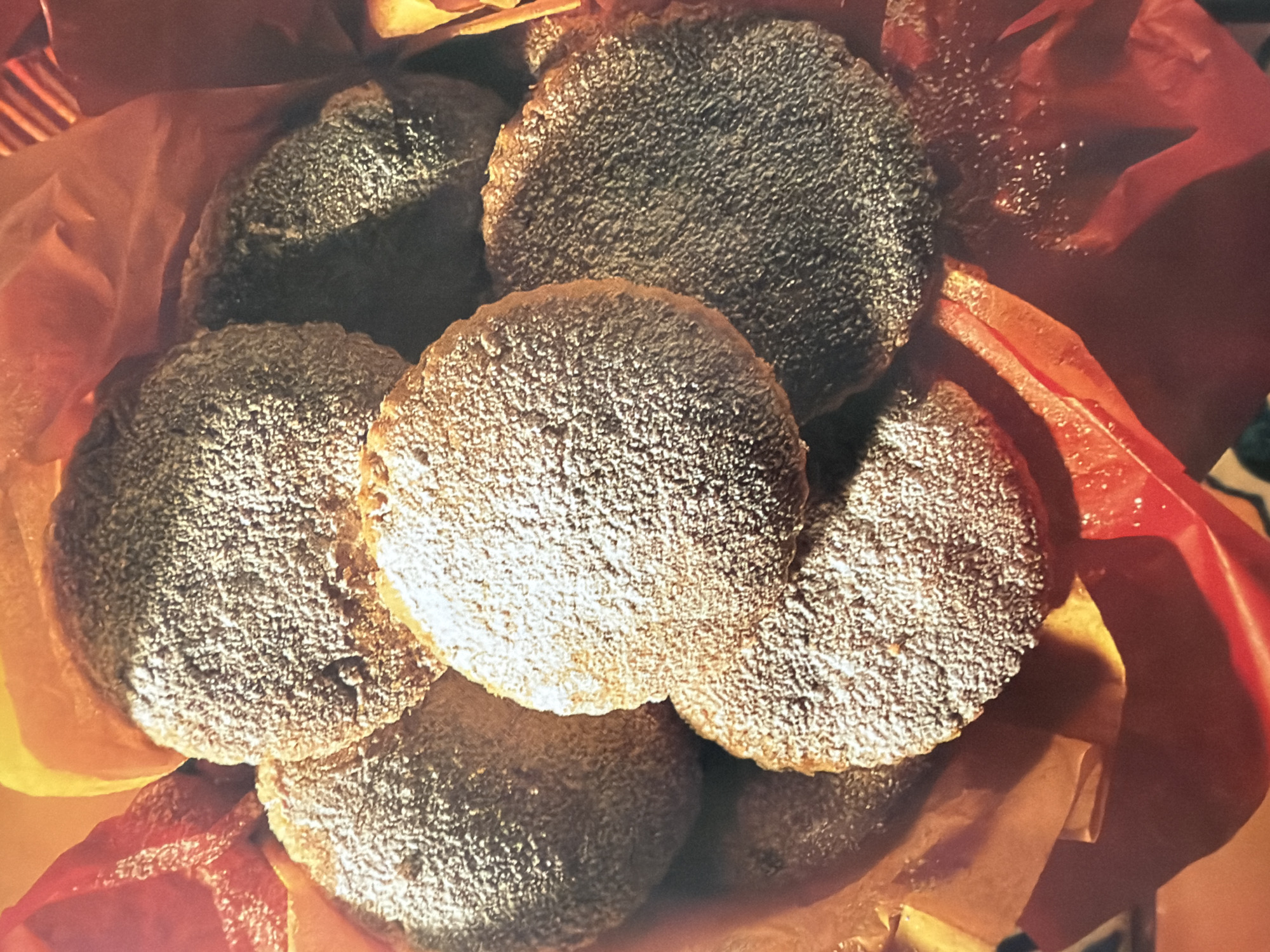 A close-up, overhead shot shows a pile of round, golden-brown cookies dusted with white powdered sugar, arranged in a red and orange crinkled paper liner, possibly from a gift basket. The cookies have a slightly textured surface and appear to be piled unevenly on top of each other. The background is filled with the bright red and orange of the paper liner, creating a festive and warm atmosphere. The lighting is soft and diffused, highlighting the powdered sugar and the warm tones of the cookies and liner. There are no people or indications of a specific city or location.