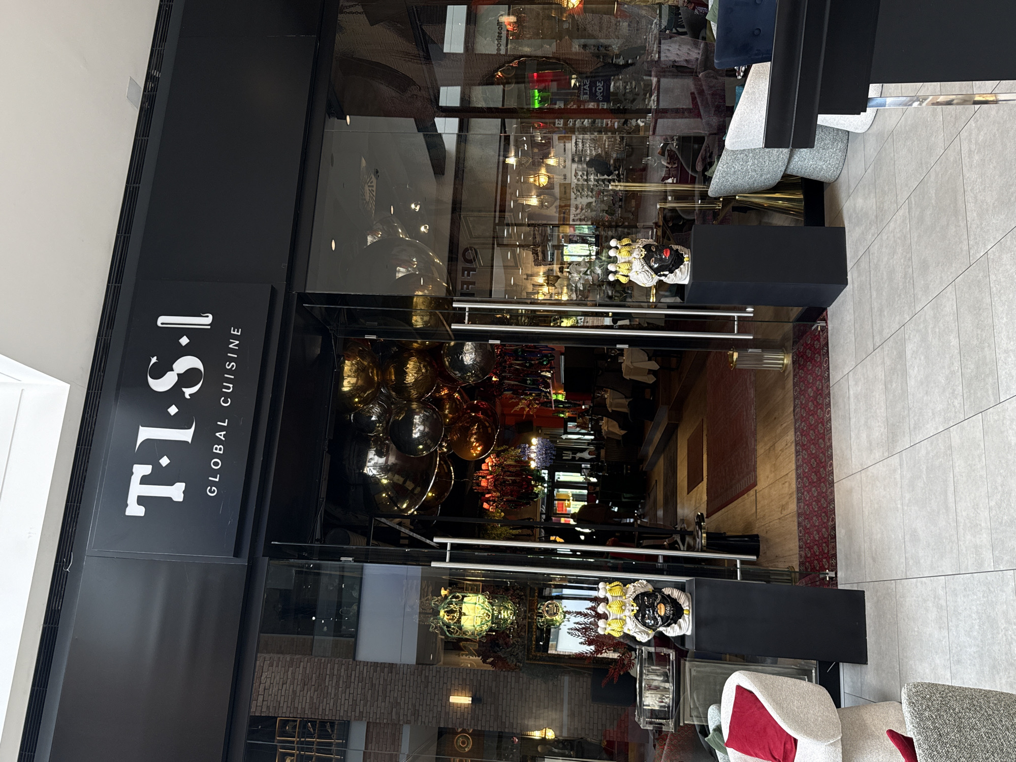 The image shows the entrance of a restaurant called "T.I.S.L Global Cuisine". It is located in Belgrade, Serbia, as evidenced by the distinctive 'Black Head' statues placed on pedestals next to the entrance. The restaurant has glass doors, and the interior is visible through the glass, revealing various decorations, light fixtures, and some seating arrangements. Outside the restaurant, the flooring consists of large, light-colored tiles. Furniture, including chairs, are also present outside the entrance. The signage on the restaurant is black with white lettering.