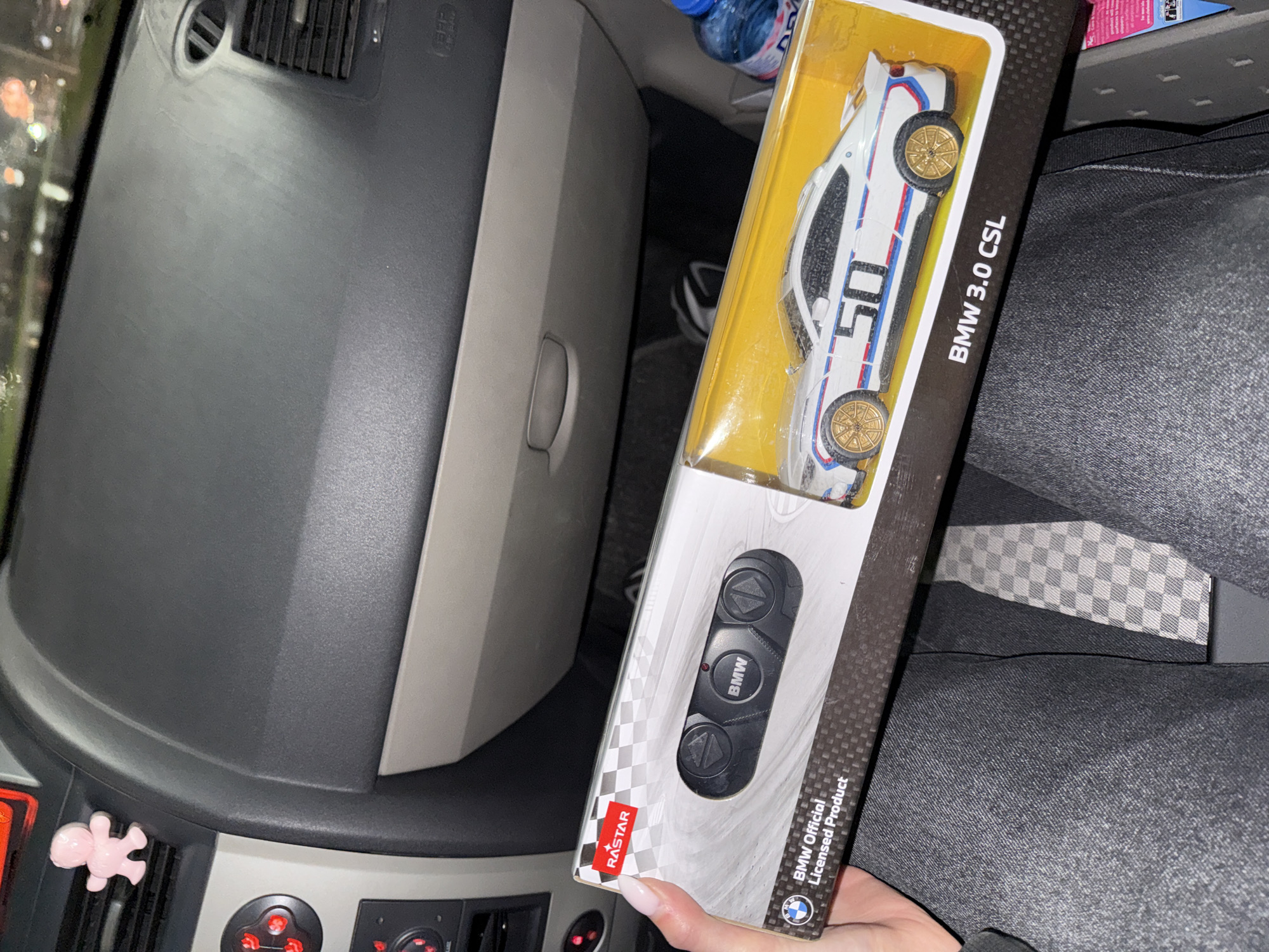 The image depicts a gray and beige car dashboard with air vents, including a pink figurine. The person holding the box of a BMW 3.0 CSL toy car is sitting in the driver seat. The location is Plovdiv, Bulgaria.