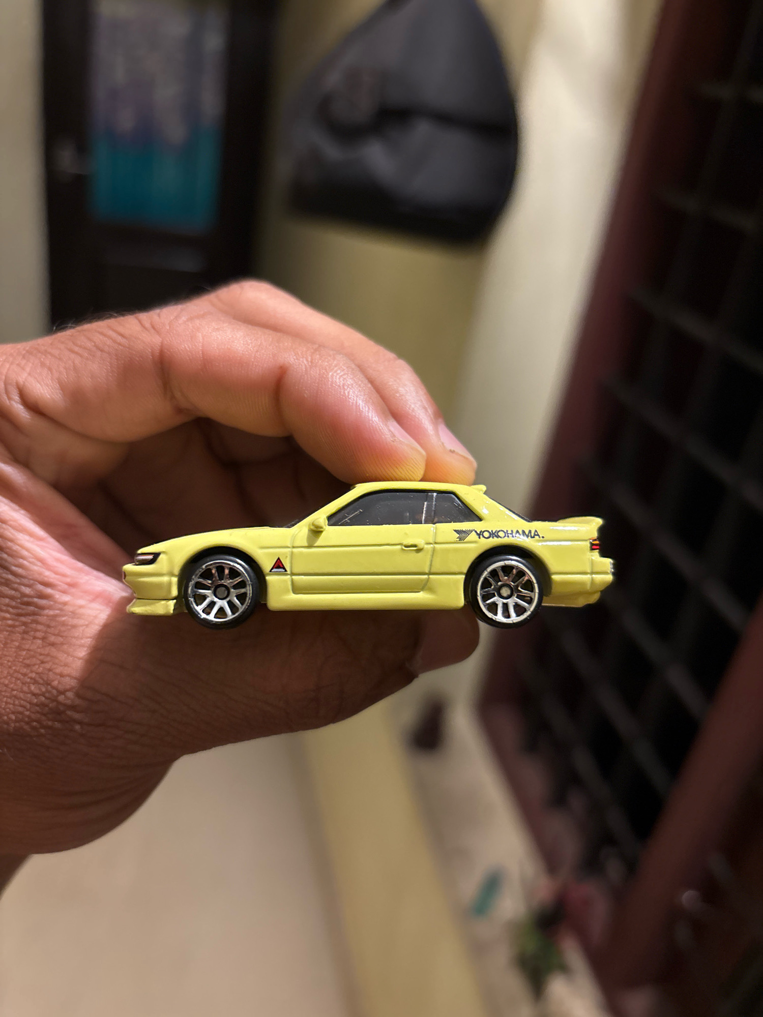 An overhead, close-up shot captures a person's hand holding a yellow toy car. The car, a miniature model, is held between the thumb and forefinger, allowing a clear view of its side profile. The toy car exhibits intricate details, including realistic wheels with silver rims and a black tire. The car's body is adorned with "YOKOHAMA" branding in white text and a small red triangle logo on the door. The background is blurred, but hints of a dimly lit room with a dark bag hanging on the wall and a dark-barred window suggest an indoor setting, potentially in a residential area of Edathala, India.