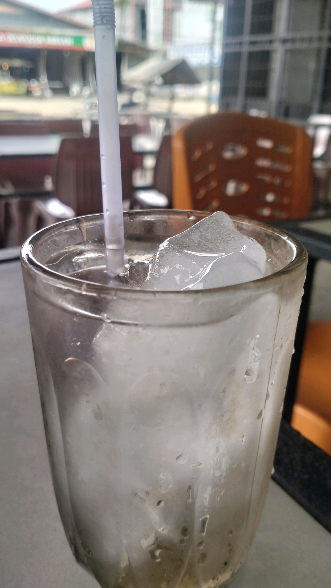 A close-up, vertical shot shows a glass of ice water with a white straw. The glass is sweating from condensation, and ice cubes fill most of its volume. The background is out of focus, but appears to be an outdoor seating area, possibly a cafe or restaurant, with chairs and tables visible. In the distance, buildings and a street are faintly discernible. The scene is likely in Lhokseumawe, Indonesia, suggested by the presence of local signage and building styles typical of the region.