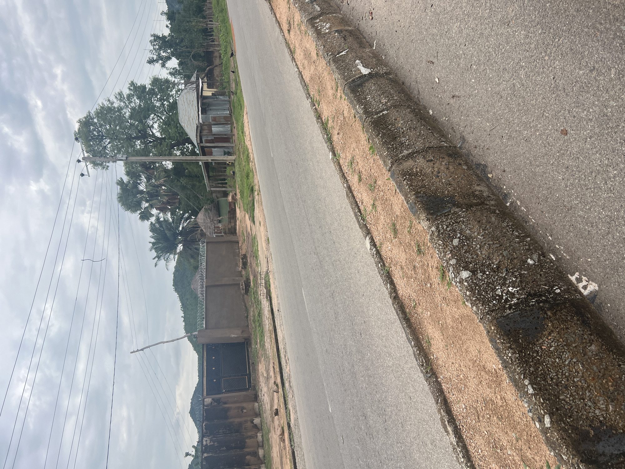 The image shows a rural road in Kona Garu, Nigeria. The road is paved and has a concrete curb on the right side. The road is bordered by a sandy embankment with sparse vegetation. On the left side of the road, there are several structures, including buildings made of corrugated metal and some huts. A utility pole with electrical wires stands prominently. Lush trees, including palm trees, are visible in the background. The sky is overcast with clouds. The image appears to be taken during daylight. There is no visible text. The scene depicts a typical rural setting with a focus on infrastructure and local architecture.