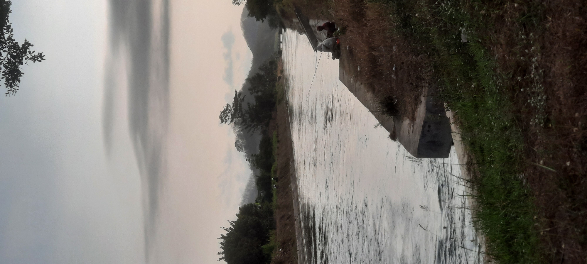 The image depicts a serene canal scene in Lodoyo, Indonesia. A wide canal, with water reflecting the overcast sky, dominates the foreground and midground. The canal's edges are lined with concrete structures and grassy banks. On the right bank, two individuals are seen fishing. One, wearing a white hooded jacket, sits with a fishing rod. Another person, in a darker garment, sits nearby. Beyond the canal, a line of trees and vegetation marks the distant bank, leading to rolling hills and mountains shrouded in mist. The sky is a soft, pale gray, with diffused clouds suggesting either early morning or late afternoon. A few branches of a tree with dark green leaves hang from the top edge of the frame, adding a natural element to the composition. The overall atmosphere is tranquil and quiet.