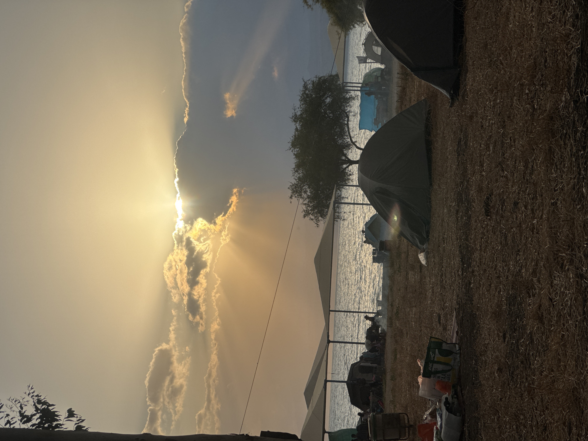 The image depicts a camping scene. Several tents are set up on dry grassy land. In the background is a body of water, possibly a lake or sea, with a few more objects visible along the shoreline. The sky is partially obscured by clouds, with the sun breaking through, creating a bright, diffused light. The location appears to be outdoors, near a body of water, with camping equipment present, likely in the city of Unknown, Unknown.