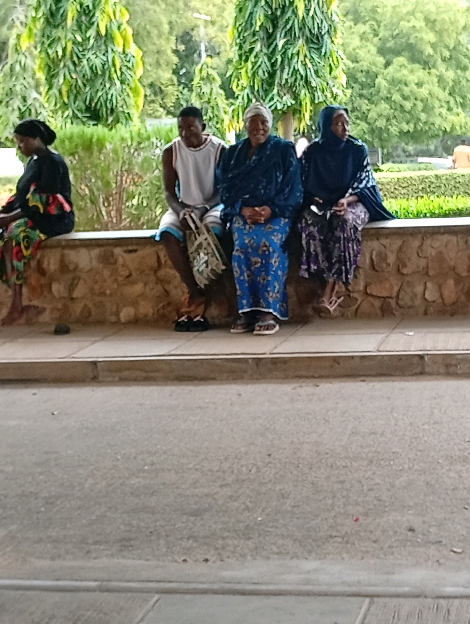 In Gombe, Nigeria, four individuals are seated on a low stone wall outdoors. From left to right, a woman in a black top and a colorful patterned skirt is seated. Next to her, a man in a white tank top and shorts is seated. He is holding a woven bag. To his right, an older woman in a blue patterned dress and headscarf is seated, her hands clasped in her lap. The final individual, a woman in a dark blue headscarf and a purple patterned dress, is seated to the right of the older woman. The wall is constructed from irregularly shaped stones and is topped with a lighter colored surface. Behind the wall, a lush green hedge is visible, backed by trees with light green foliage. In the foreground, a paved surface, likely a road or walkway, is present, with scattered debris.