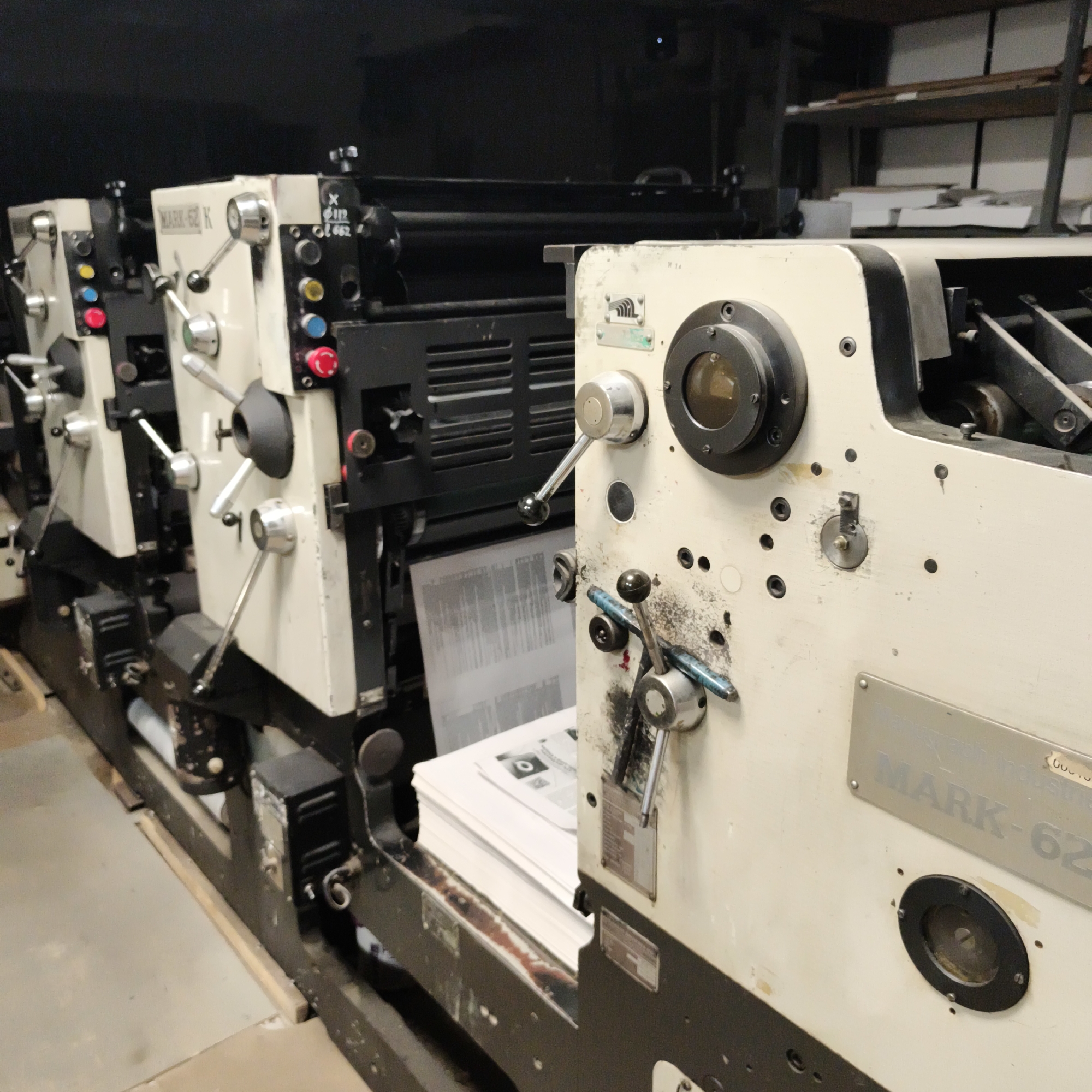 The image shows a close-up view of the side of a printing press, likely in an industrial setting. The machine is primarily cream-colored with black accents and various metal levers, knobs, and small windows. One prominent feature is a large circular glass viewer on the right side of the machine. There are control buttons with colored lights (red, yellow, blue) and other indicators on the left side of the press. Stacks of white paper are visible in the lower part of the image, suggesting the press is in operation or ready to print. The background is dark and slightly out of focus, with shelves containing what appear to be paper or other materials visible in the upper right corner. The overall impression is one of a functional, but potentially aged, industrial printing machine. The location in Chaikovsky, Russia, is a matter of fact from the query and not visually discernible from the image itself.