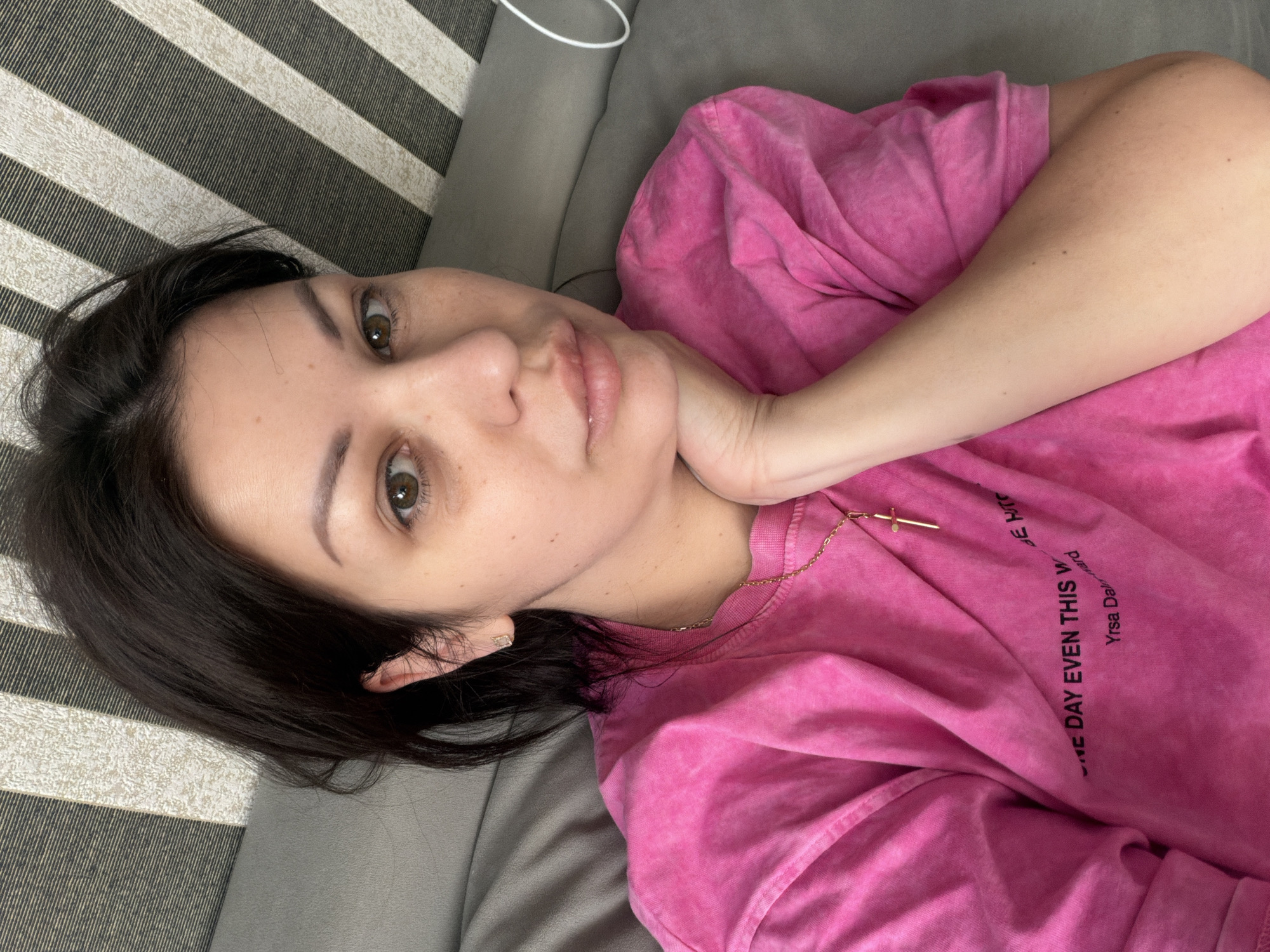 A woman with dark hair and brown eyes is lying on a gray couch. She is wearing a pink t-shirt with black text that reads "ONE DAY EVEN THIS WARD Yrsa Dalm". A gold cross necklace is visible on her chest. Her left hand is resting on her chin, and her right arm is bent. The background consists of a striped headboard, with alternating black and white stripes. The scene appears to be indoors, likely in a bedroom or living room. There is no information to indicate this image is from Sochi, Russia.