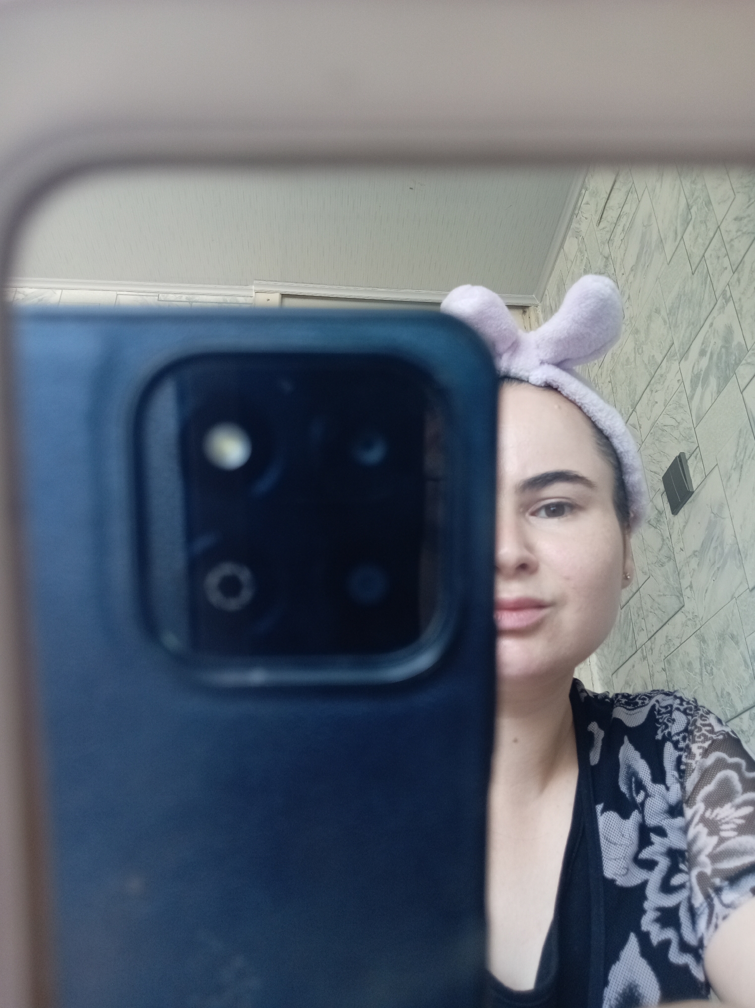 A woman is taking a selfie in a mirror. She is wearing a light purple headband with bunny ears. Her right eye, nose, and mouth are visible. She is wearing a black top with a grey and black floral print cardigan over it. The background shows a tiled wall and a white ceiling. A dark-colored smartphone is held up, obscuring the left side of her face and the mirror's edge. The scene is likely indoors, possibly in a bathroom or bedroom in Khujand, Tajikistan.