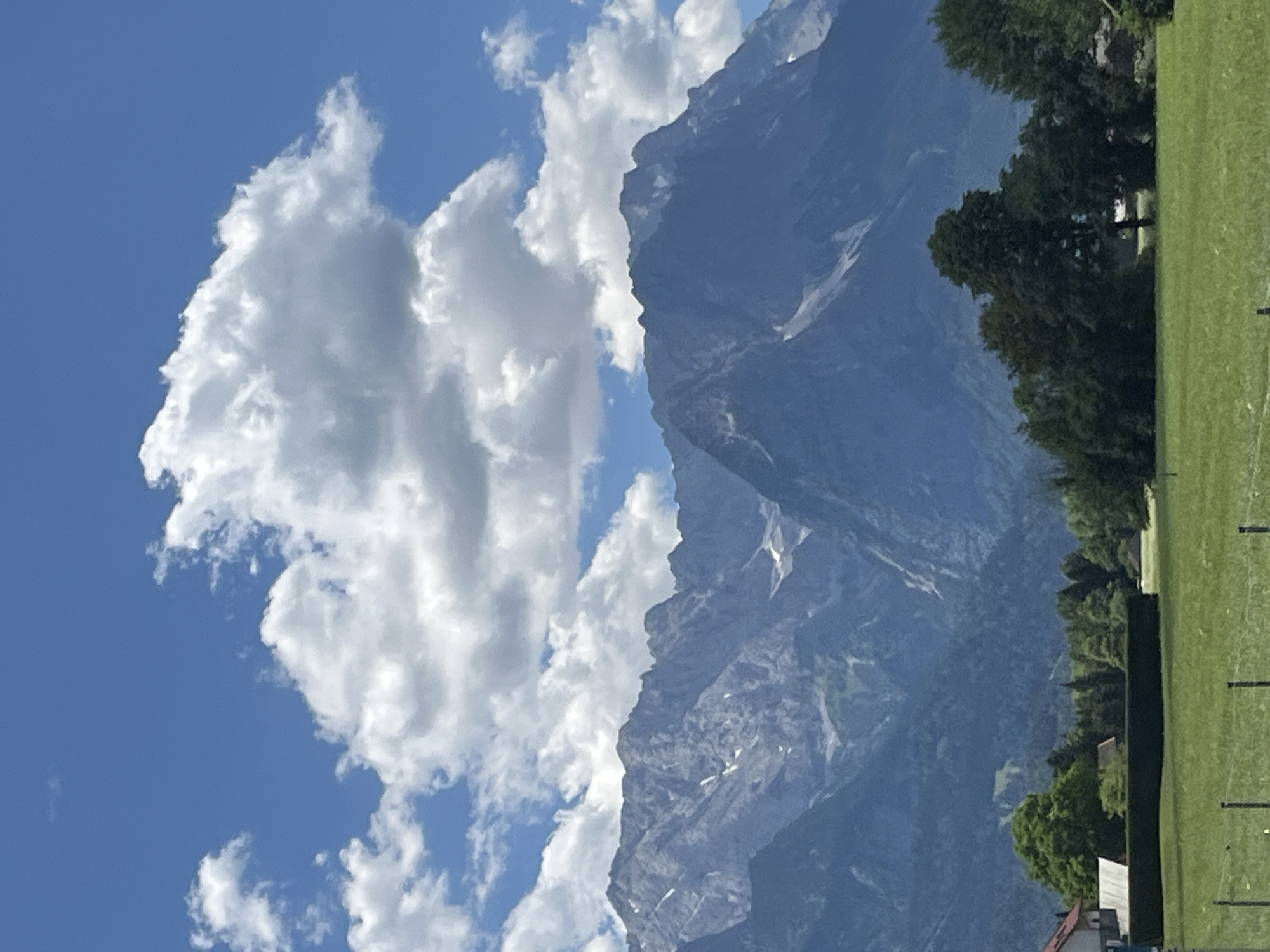 The image shows a landscape with mountains in the background, a green field in the foreground, and some trees and buildings. The sky is blue with white clouds. Specifically, the image appears to capture a view of the Zugspitze massif, Germany's highest peak, located in the Wetterstein mountain range. The presence of the Zugspitze and the surrounding Bavarian Alps strongly suggests the location is near Garmisch-Partenkirchen, Germany. The foreground shows a well-maintained green lawn, possibly a park or agricultural field, bordered by a dark green hedge. To the left, a red-roofed building with white walls is partially visible, suggesting a residential area. Several trees, both deciduous and coniferous, are scattered across the scene, with denser forestation further back towards the mountains. The mountains themselves are rugged and rocky, with some patches of snow visible on their higher slopes. The clouds are fluffy and white, indicating a bright, sunny day. There are no people visible in the image. The scene is peaceful and serene, characteristic of a rural or mountainous environment.