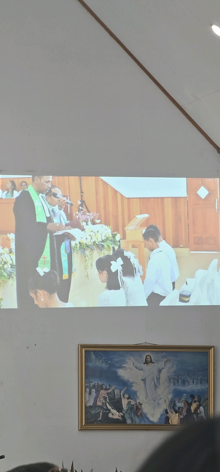 The image displays a projected image of a religious ceremony onto a wall. In the foreground of the projected image, a person wearing a black robe and a green stole stands behind a lectern, holding a book and a microphone. They are speaking or leading a service. To their right, another person in a white shirt is visible. In front of the lectern, two young females, each wearing white dresses and elaborate white bows in their hair, are seated. Behind them, a young male in a white shirt stands. The background of the projected image shows a wooden wall and a door. Below the projected image, a framed painting of a religious scene, likely the resurrection of Jesus, is hung on the wall. The overall environment suggests a place of worship or a religious gathering, possibly in Puncak Jaya, Indonesia.
