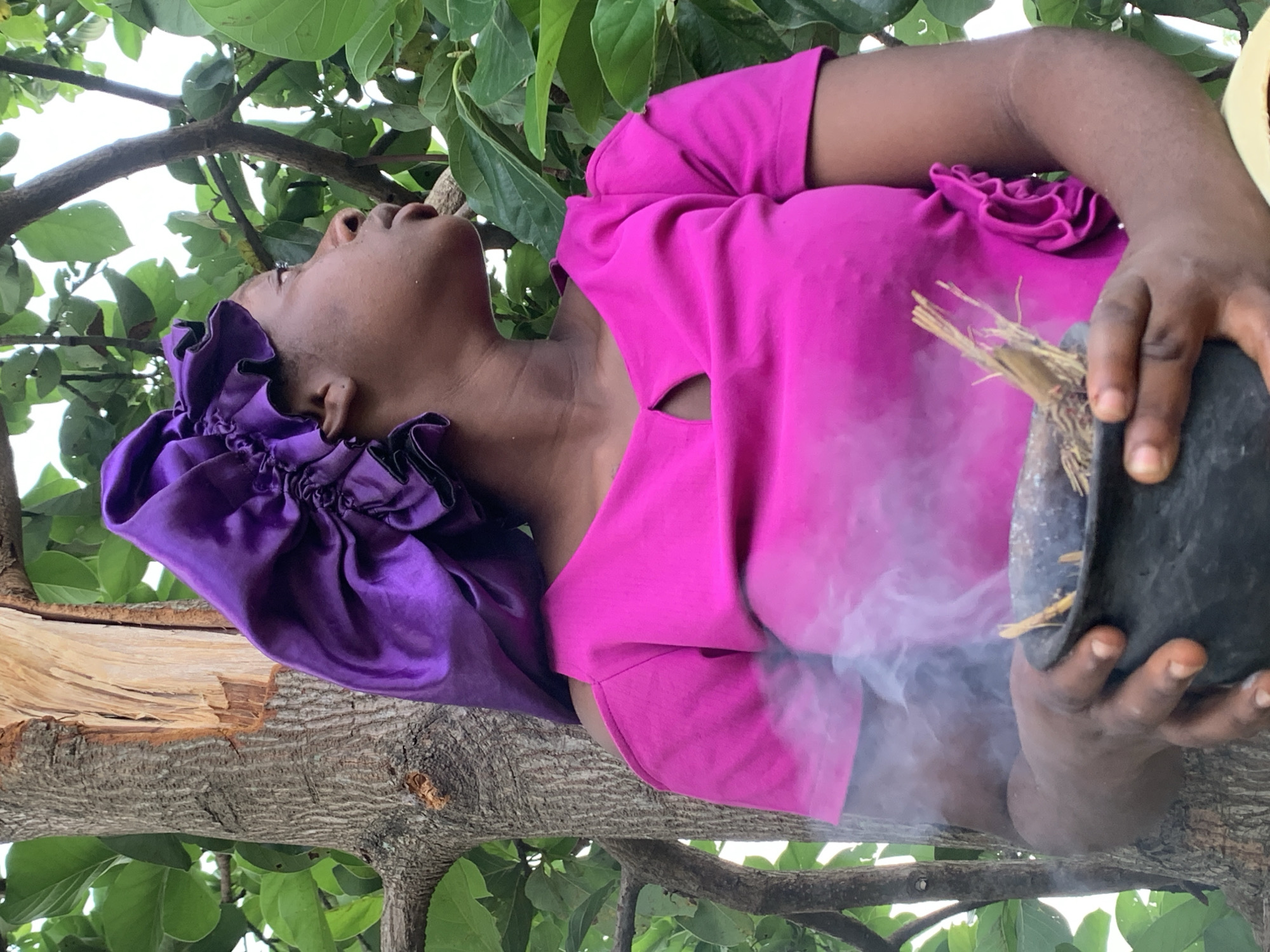 A woman is sitting on a tree branch looking up. She is wearing a bright pink shirt and a ruffled purple bonnet. She is holding a black pot containing burning herbs or incense. Smoke is wafting upwards. The setting is outdoors, with the tree branch surrounded by green leaves. The location is Agban, Nigeria.