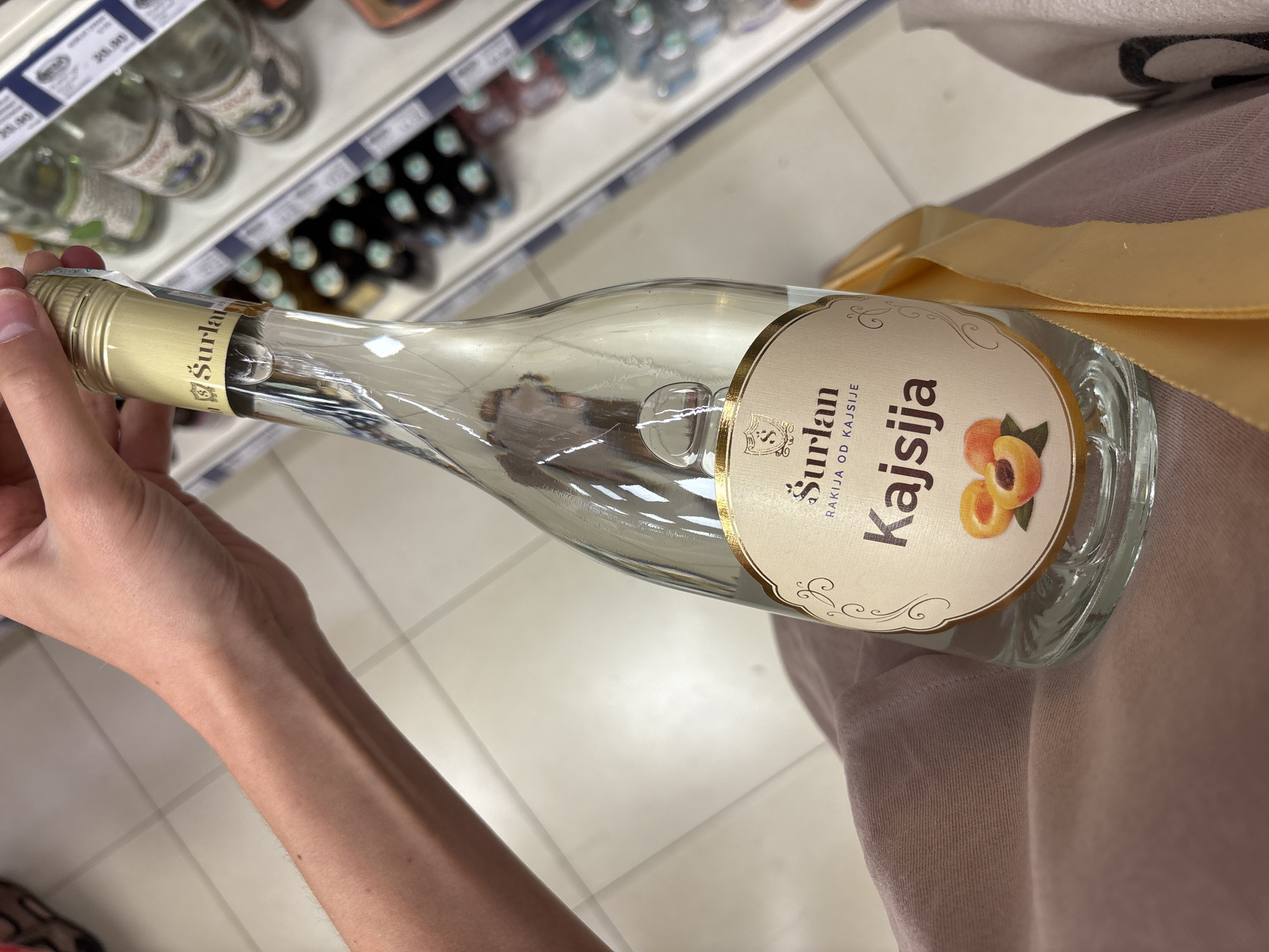 In Boreti, Montenegro, a person holds a clear glass bottle of "Šurlan Kajsija," a type of rakija made from apricots. The bottle has a gold-colored neck and a cream-colored label adorned with the brand name "Šurlan," the words "RAKIJA OD KAJSIJE," and an illustration of apricots. The background suggests a retail environment, possibly a supermarket aisle, with shelves stocked with various bottles and beverages. The person's arm and hand are visible, holding the bottle, with a light-colored fabric (likely clothing) draped in the foreground. The floor is tiled with a light-colored pattern.