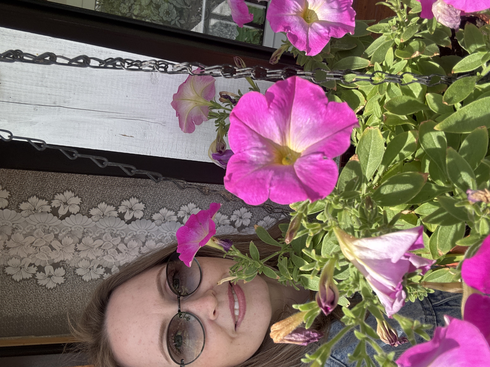 A woman with light brown hair and round, wire-rimmed sunglasses is positioned to the left of a cluster of bright pink petunia flowers and green foliage. A metal chain hangs above the flowers, suggesting they are in a hanging planter. Behind the chain is a white wooden plank and above that is a blurred decorative pattern that is floral. A section of the woman's face, including her nose, mouth, and eyes (as visible through the sunglasses), is visible. The overall impression is of a close-up, possibly a selfie, taken outdoors. The location is unknown, but the request includes Chaikovsky, Russia.