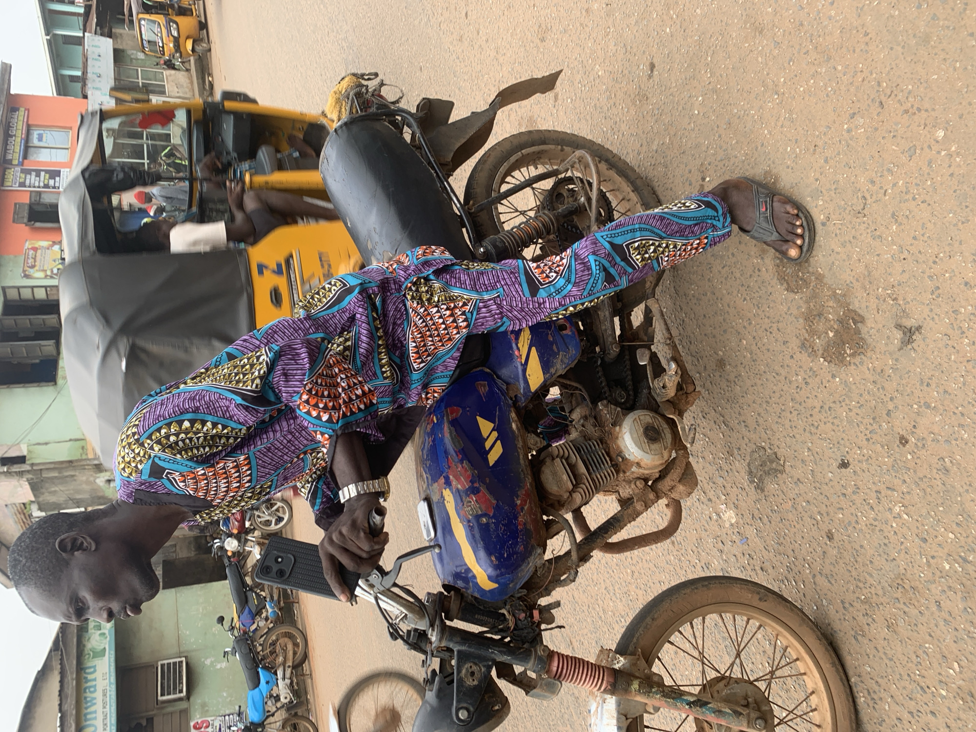 A man wearing a colorful African print outfit is sitting on a blue and yellow motorcycle on a street in Shagamu, Nigeria. The motorcycle is muddy and appears to be well-used. In the background, a yellow auto-rickshaw, also known as a Keke Napep, is visible, as are other motorcycles and buildings lining the street. The ground is paved with asphalt and is covered in dirt and debris. The sky is overcast, suggesting a cloudy day. The man is holding the handlebars of the motorcycle and looking forward.