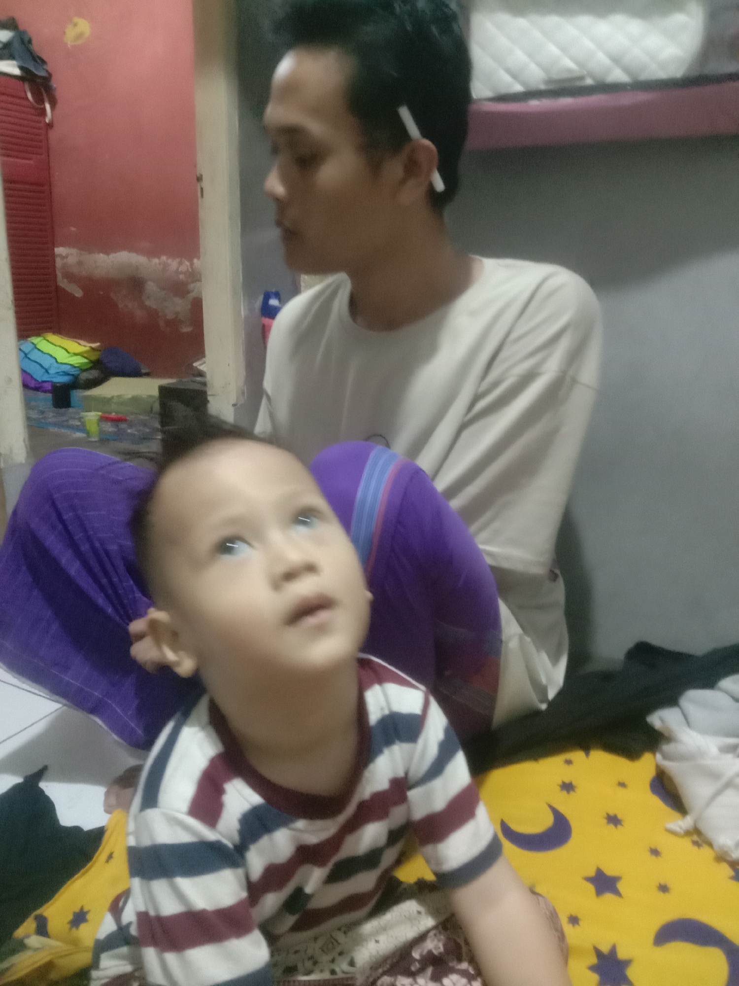 The image captures a close-up view of two individuals, likely a father and son, in an indoor setting. The child, positioned in the foreground, is wearing a striped shirt and looking upwards. The adult, in the background, is wearing a light-colored t-shirt and has a cigarette tucked behind his ear. The environment appears to be a room with a red wall and some scattered items, including what looks like a colorful mat and fabric. The overall impression is a candid snapshot of a domestic scene.