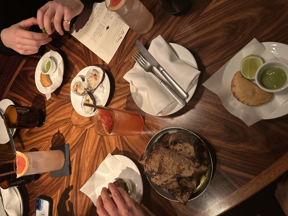An overhead shot captures a dining scene at a restaurant in London, United Kingdom, likely in the evening given the warm, ambient lighting. Several hands are visible around a dark, highly polished wooden table with a striking starburst grain pattern, suggesting multiple people are sharing a meal.
The table is laden with various appetizers and drinks. In the upper left, a person's hands are seen squeezing a lime wedge, possibly over a dish. Nearby is a small white plate holding a single fried pastry (possibly an empanada) and a small bowl of vibrant green dipping sauce. To the right, a plate of three fresh oysters on a bed of ice is presented, garnished with what appears to be red and green roe or small diced vegetables.
Further right, a white plate with a neatly folded white napkin holds a fork and knife, ready for a diner. On the opposite side of the table, a metal bowl contains several slices of grilled bread, glistening with oil. Numerous cocktails in tall glasses, garnished with slices of grapefruit, are scattered across the table, some with straws. Darker glasses, possibly for water, are also present. One person's hand is visible near another small bowl of green dipping sauce. A smartphone in a clear case with small cherry graphics is also visible on the table.
A menu titled "La Musa" is prominently displayed near the top, indicating the establishment's name. The overall impression is one of a sophisticated and social dining experience, with a focus on shared plates and craft cocktails.