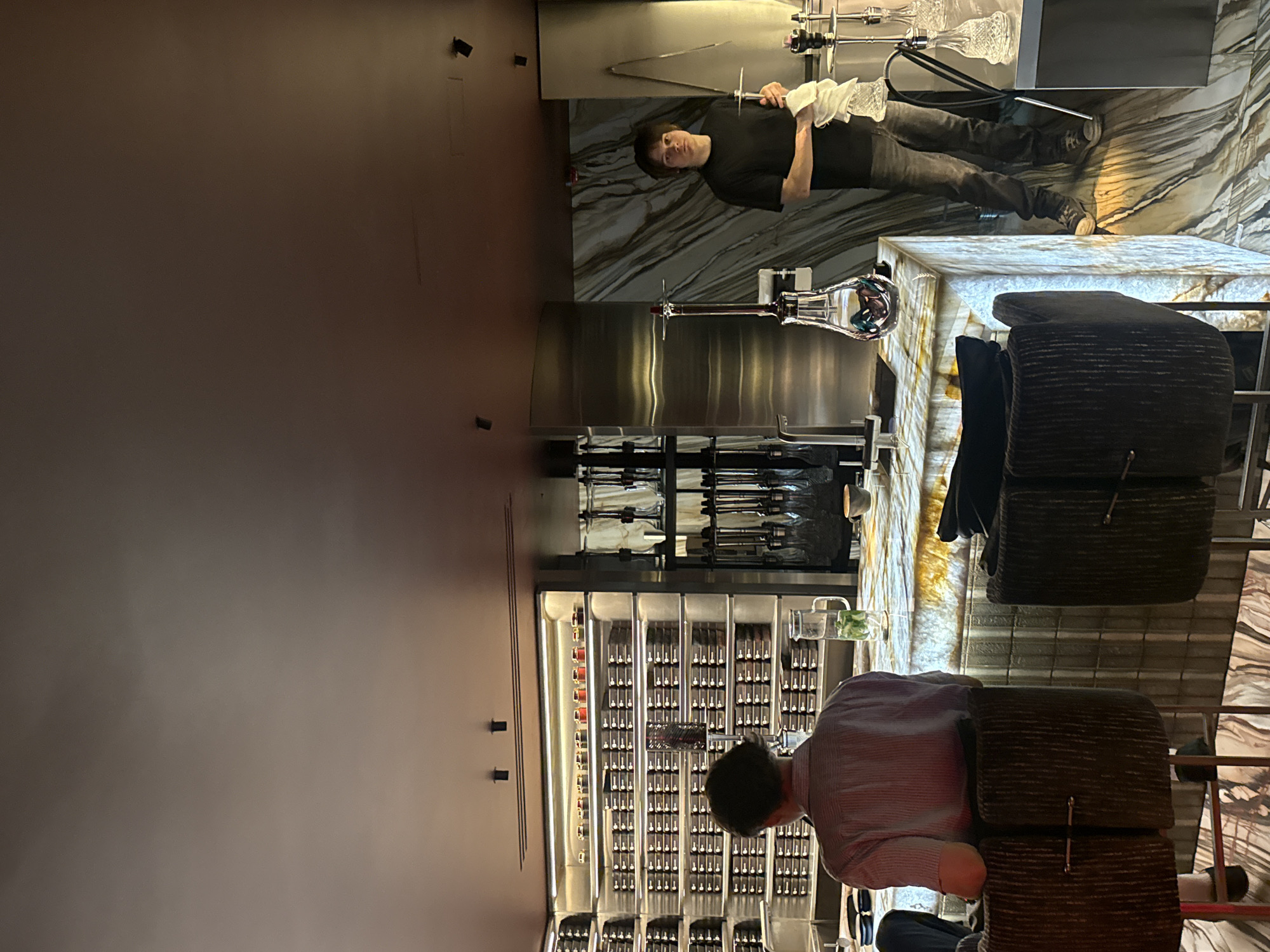 This image captures the interior of a modern, upscale lounge or bar, likely during the evening. The setting is characterized by sophisticated design and ambient lighting.
In the right portion of the frame, a man in a dark t-shirt and pants stands behind a dark, veined marble counter. He holds a white cloth and a slender metal instrument, appearing to prepare or attend to one of the several ornate glass shishas or hookahs displayed prominently on the counter and shelving behind him. These shishas feature intricate glass components.
In the foreground and central-left, another man, viewed from behind, is seated in a dark, textured armchair at a luminous, translucent bar or table. He is wearing a striped shirt and is looking towards a large, illuminated wall display. This display is composed of numerous small, uniformly arranged bottles or vials within a grid system, suggesting a vast collection of spirits, mixers, or shisha flavors.
The flooring throughout the visible area is a dark, patterned marble, complementing the main counter. The walls are a muted, dark brown, enhancing the refined atmosphere. Several small, black, rectangular fixtures are mounted on the walls. The overall scene suggests a calm, luxurious environment for social gatherings or relaxation. No discernible text is visible in the image.