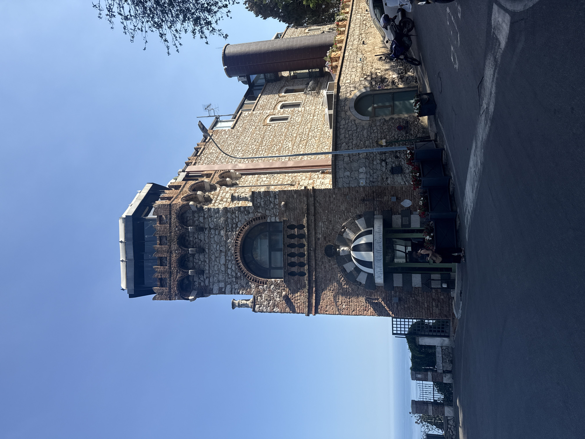 This image captures the Hotel Villa Carlotta in Taormina, Italy, on a clear, sunny day. The prominent structure is a multi-story building constructed with a mix of rough-hewn stone and reddish-brown brickwork, exhibiting a fortified, castle-like architectural style. The leftmost section features a tower-like element with crenellated brickwork supporting a modern, glass-enclosed observation deck at its apex. Below this, a large arched window with a small balcony is visible. A decorative white bust is positioned on a stone ledge to the left of the arched window. The hotel's main entrance is situated beneath a black and white striped domed awning, with "Hotel Villa Carlotta" inscribed above it.
To the right of the entrance, the building continues with predominantly stone walls, featuring multiple rectangular windows and a tall, dark, cylindrical structure. A grey street light pole stands in front of this section of the building. The scene is set on an asphalt road that curves gently in the foreground. A lone individual, dressed in dark attire and holding a bag, stands beneath the striped awning near the hotel entrance, appearing to be looking down. Parked on the right side of the road, in front of a lower stone wall adorned with flower planters, is a blue Yamaha motorcycle. Parts of a white car and another motorcycle are partially visible further to the right. In the far background, to the left, the calm blue waters of the Mediterranean Sea are visible under a clear, cloudless sky. A black metal fence with stone pillars separates the road from the drop-off towards the sea. Some dark green foliage is visible on the far right.