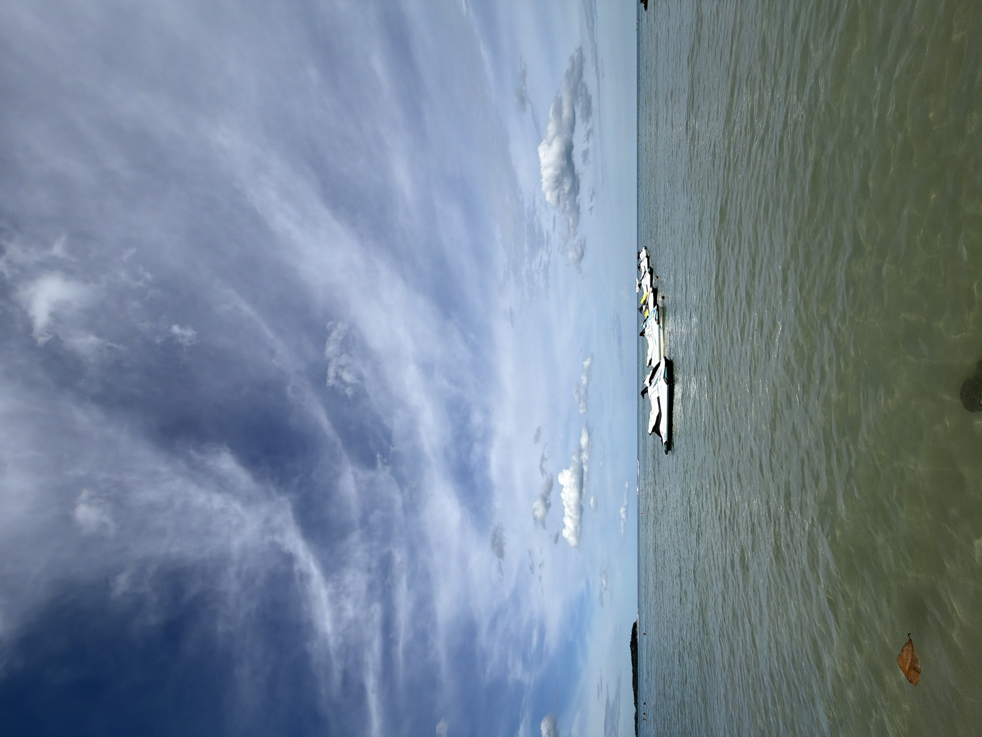 A serene, wide-angle shot captures the coastline of Baan Chaweng, Thailand, under a vast, cloudy sky. The foreground is dominated by calm, greenish-brown water, with a single dry leaf floating near the bottom left corner, hinting at the gentle lapping of waves. In the mid-ground, a line of several white and yellow jet skis are moored in a row, bobbing gently on the water. Further out, the horizon is marked by the faint silhouette of a lush green island with a few scattered people visible on its shore. The sky above is a dynamic canvas of blues and whites, with wispy, elongated clouds stretching across the frame, creating a sense of depth and atmospheric movement. The overall scene conveys tranquility and the inviting calm of a tropical seascape.