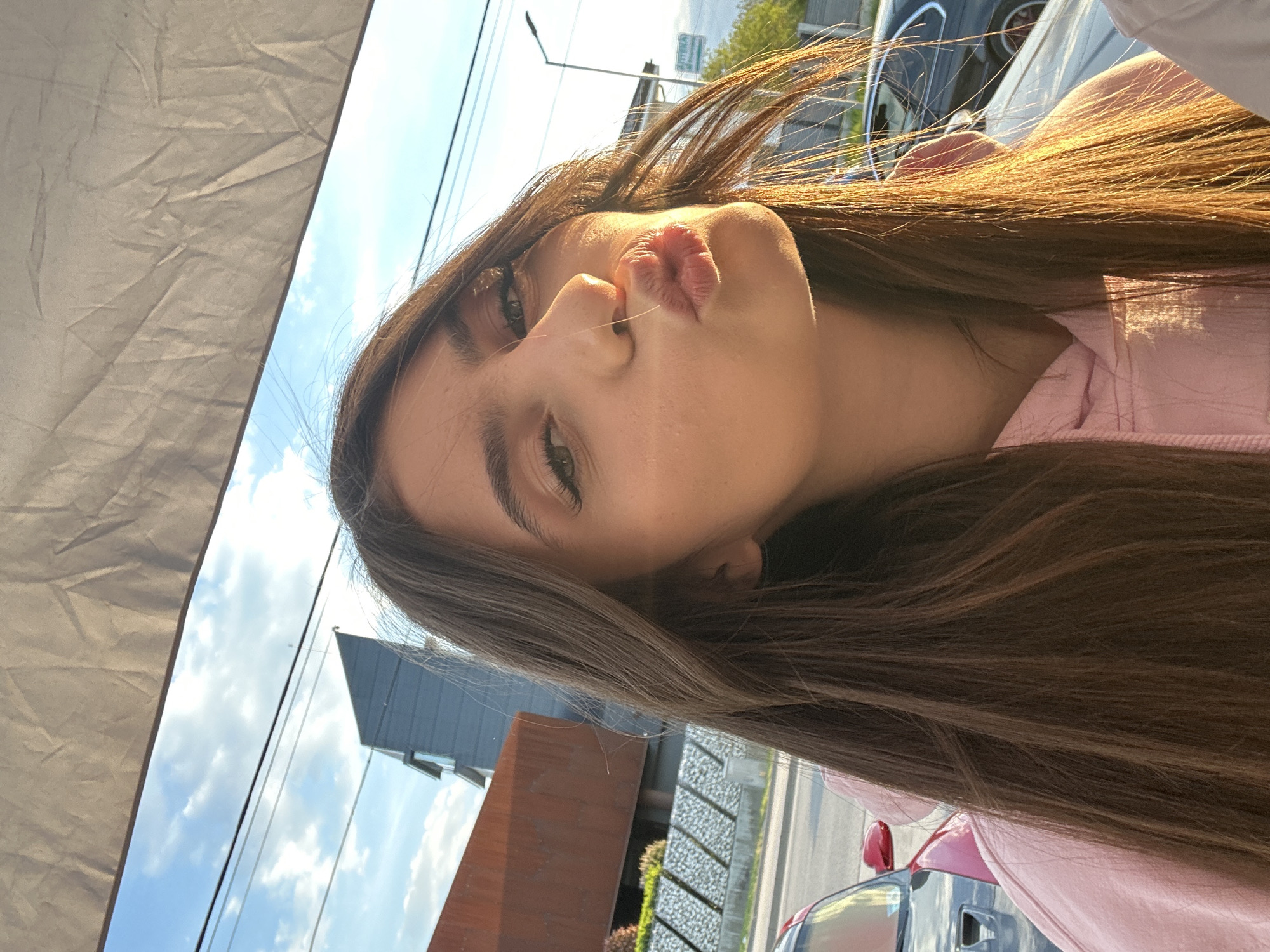The image captures a close-up, rotated portrait of a young woman taken outdoors, likely in Plovdiv, Bulgaria. She is the central subject, with her head and upper torso visible, and her long, straight brown hair with sunlit, reddish highlights flowing across the frame. Her lips are pursed in a playful "kiss" or "duck face" expression, and her eyes are open, looking towards the camera. She appears to be wearing a pink top or hoodie.
The lighting suggests late afternoon or early morning, with warm, golden sunlight illuminating her hair and face, casting some soft shadows. The sky in the background is bright blue with scattered white clouds. Various elements of an urban environment are visible behind her. A light-colored canvas or fabric, possibly an awning or umbrella, occupies the upper left of the frame. In the mid-ground, a street light pole stands against the sky, alongside what appears to be a modern building with reflective surfaces and another structure with a distinctive rusty brown facade. A car, partially red and black, is visible at the bottom right. The overall impression is a casual, cheerful moment captured in bright daylight. No discernible text is visible in the image.
