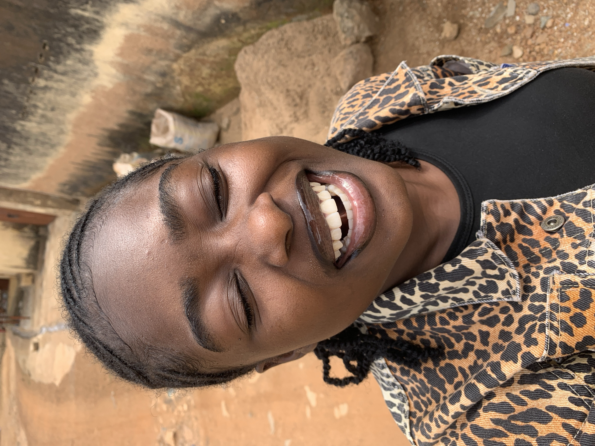 A close-up, slightly high-angle portrait features a young Black female in an outdoor setting in Rafin Par, Nigeria. She is observed with her eyes closed and her mouth open in a wide smile, revealing white teeth and glossy, dark lips. Her hair is styled in cornrows along the scalp. She wears a black garment visible at the neckline, beneath a collared jacket patterned with a leopard print featuring tones of tan, black, and off-white.
The background consists of a reddish-brown dirt ground, consistent with an unpaved road or path, receding into the distance on the left. A narrow channel, possibly for water runoff, is visible along the left side of the path. To the right, the terrain features multiple large, irregularly shaped brown rocks. A weathered wall with vertical and horizontal striations of muted brown and beige tones is partially visible in the middle ground, behind which a white sack, possibly containing granular material, is discernable. The scene is illuminated by natural daylight.