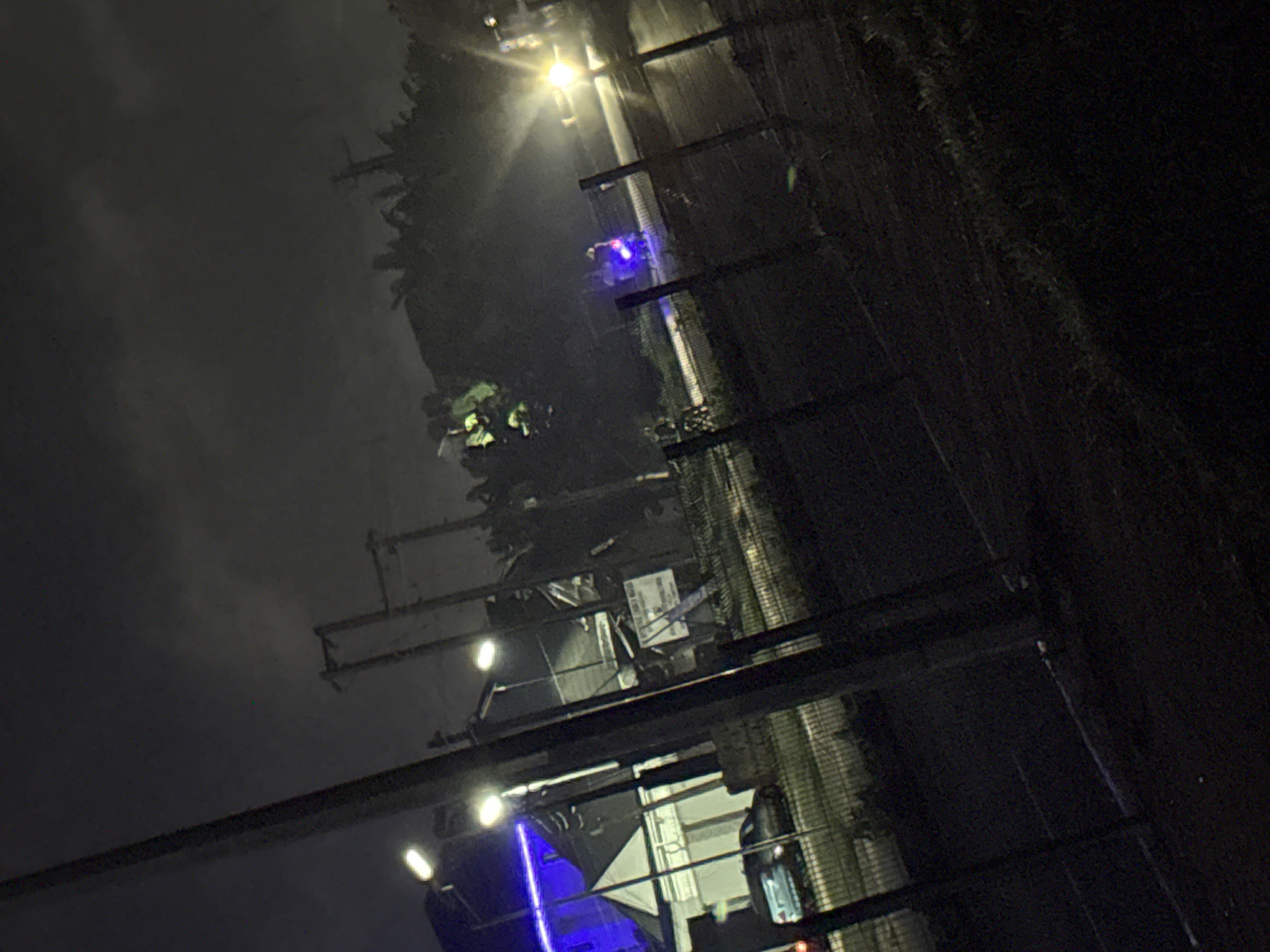 The image depicts a nighttime scene on a street in Uyo, Nigeria. A dark road with a chain-link fence in the foreground dominates the lower half of the frame. Beyond the fence, a car is parked on the side of the road, and a building with bright blue lights on its facade is visible. Further down the road, a person on a motorcycle with illuminated blue lights is seen. Streetlights and a bright vehicle headlight illuminate parts of the scene, contrasting with the overall darkness and cloudy sky. The environment appears to be urban or suburban, with indications of infrastructure like utility poles.