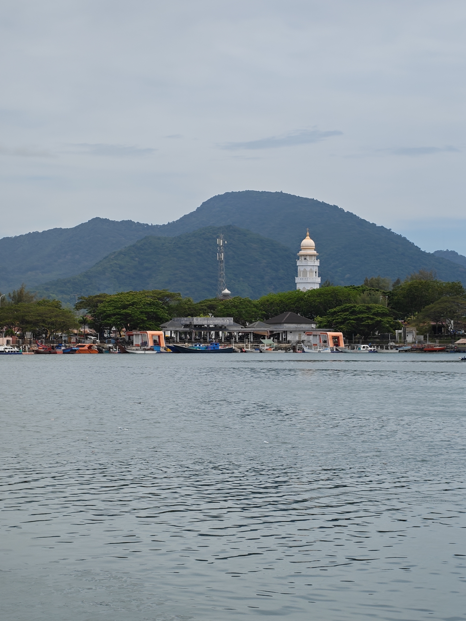 The image depicts a coastal scene in Banda Aceh, Indonesia. The foreground is dominated by a body of water, with gentle ripples visible on its surface. In the middle ground, a coastline is lined with various boats and structures, including what appears to be a small building with an orange exterior, along with an open structure, suggesting a harbor or docking area. In the background, a prominent mountain range provides a scenic backdrop, with a tall, white minaret with a golden dome rising amidst the trees, indicating a mosque. A communication tower is also visible near the mountains. The sky is overcast.
