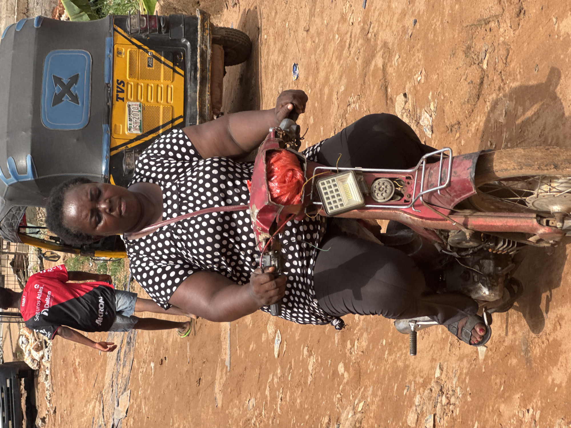 A woman is sitting on a red motorbike, positioned at an angle in the image's center. She's wearing a black and white polka dot shirt and black pants. The motorbike appears old and slightly dilapidated. A yellow and black TVS tricycle is partially visible to the left of the woman. A person wearing a red and black shirt is walking to the left of the motorbike. The ground is a mix of dirt and what seems to be a paved or semi-paved surface. The image was taken during daylight, with the lighting creating shadows. This scene is located in Onitsha, Nigeria.