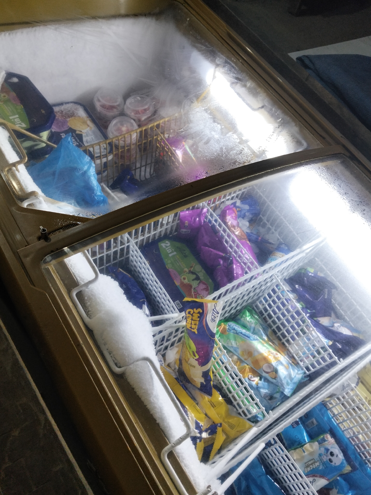 This is a close-up, high-angle, indoor shot of a freezer chest filled with various ice cream products. The freezer has a glass lid, which is partially covered in condensation and frost, obscuring the contents. Inside, multiple white plastic baskets hold an assortment of individually wrapped ice cream bars and small tubs of ice cream. Some of the packaging is brightly colored, featuring images of fruits and characters. There is a visible layer of frost and accumulated ice on the interior surfaces and some of the products. In the upper left section, a blue plastic bag is visible, and in the foreground, on the left edge of the freezer, a portion of a metal handle is covered in snow. The lighting appears to be coming from within the freezer, highlighting the products and the frosty environment. This image is consistent with the interior of a commercial freezer found in a convenience store or small grocery in Davao City, Philippines, given the type of products typically sold.
