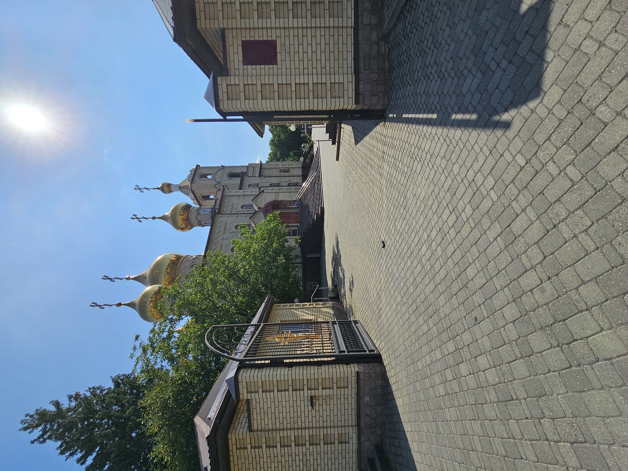 The image captures the entrance to a church in Cherkessk, Russia, on a bright, sunny day. The foreground is dominated by a paved courtyard made of grey cobblestones. To the left, a gate with intricate metalwork, featuring a golden cross, stands open, leading to a brick building with a dark-tiled roof. To the right, another brick building with a similar roof and a dark red plaque is visible.
In the background, a large, light-colored church with multiple golden domes topped with crosses stands prominently against a clear blue sky. The sun is shining brightly, casting shadows across the courtyard. Lush green trees are scattered around the church and behind the buildings. The architecture suggests a Russian Orthodox church. There are no people visible in the image.