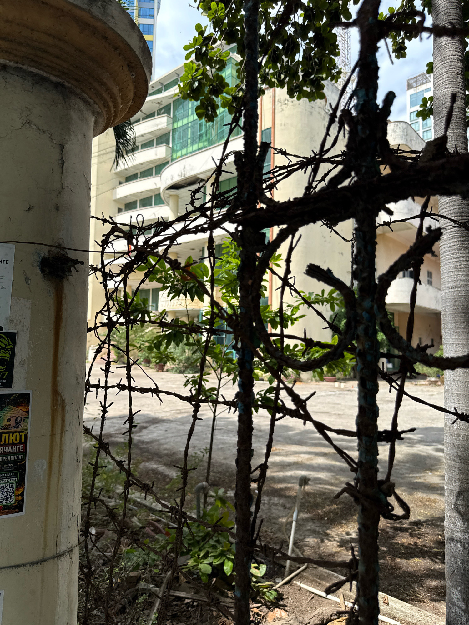 This image captures a close-up view of a weathered, cream-colored pillar and a rusty barbed wire fence in Nha Trang, Vietnam. The pillar, showing signs of age with peeling paint and streaks of dirt, is partially obscured by the barbed wire. Through the fence and foliage, a modern building with multiple balconies and green-tinted windows is visible, suggesting an urban setting. The ground appears to be a paved area or courtyard. A palm tree trunk is visible on the right side of the frame, further indicating a tropical or subtropical location. The sky above is overcast with some patches of blue.