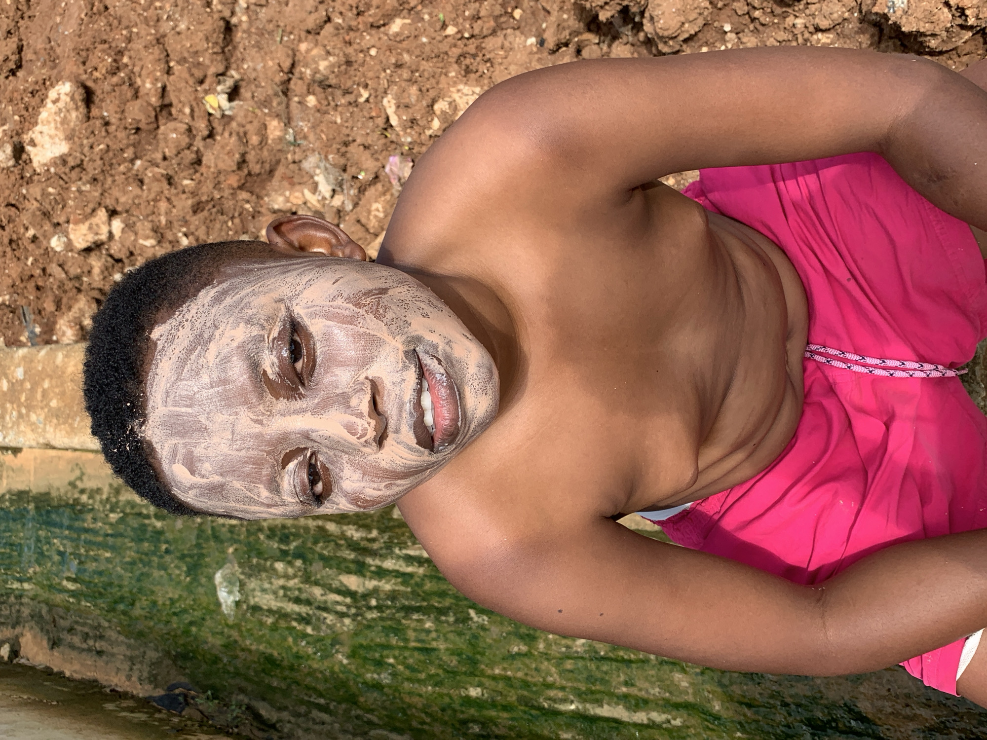 This is a portrait of a young Black boy wearing bright pink shorts. He has a light-colored face mask applied to his face, covering his forehead, cheeks, nose, and mouth. His eyes are open, and he appears to be smiling slightly, revealing his teeth. The boy is positioned outdoors, with a background of dry, reddish-brown soil with some stones and debris. In the foreground, there is a body of greenish water with some moss or algae. The lighting suggests it is daytime. The overall scene seems to be in a natural or semi-natural outdoor setting, possibly near a body of water or a construction site given the soil. The image is taken from a slightly elevated angle, looking down at the boy.