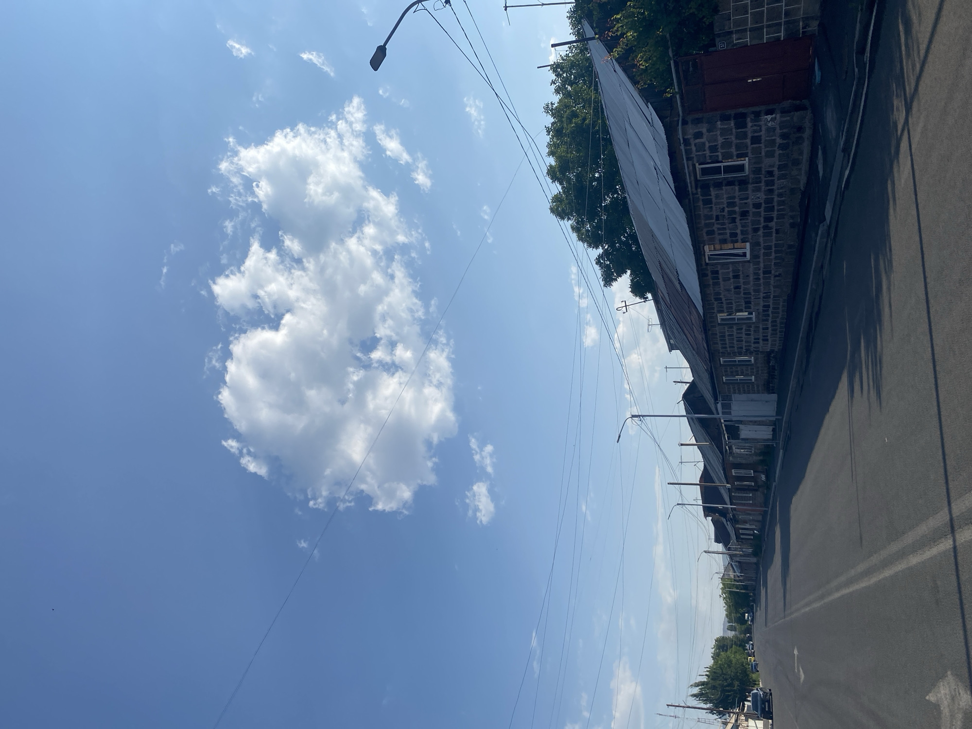 The image shows a street scene in a residential area with houses lining one side of the road. The houses appear to be older, with stone walls and corrugated metal roofs. There are trees and vegetation visible behind the houses. The street is paved and has double yellow lines down the center. There are utility poles and wires running along the street. The sky is blue with some scattered clouds. A single car is parked on the left side of the street. The overall impression is of a quiet, somewhat run-down neighborhood on a sunny day.