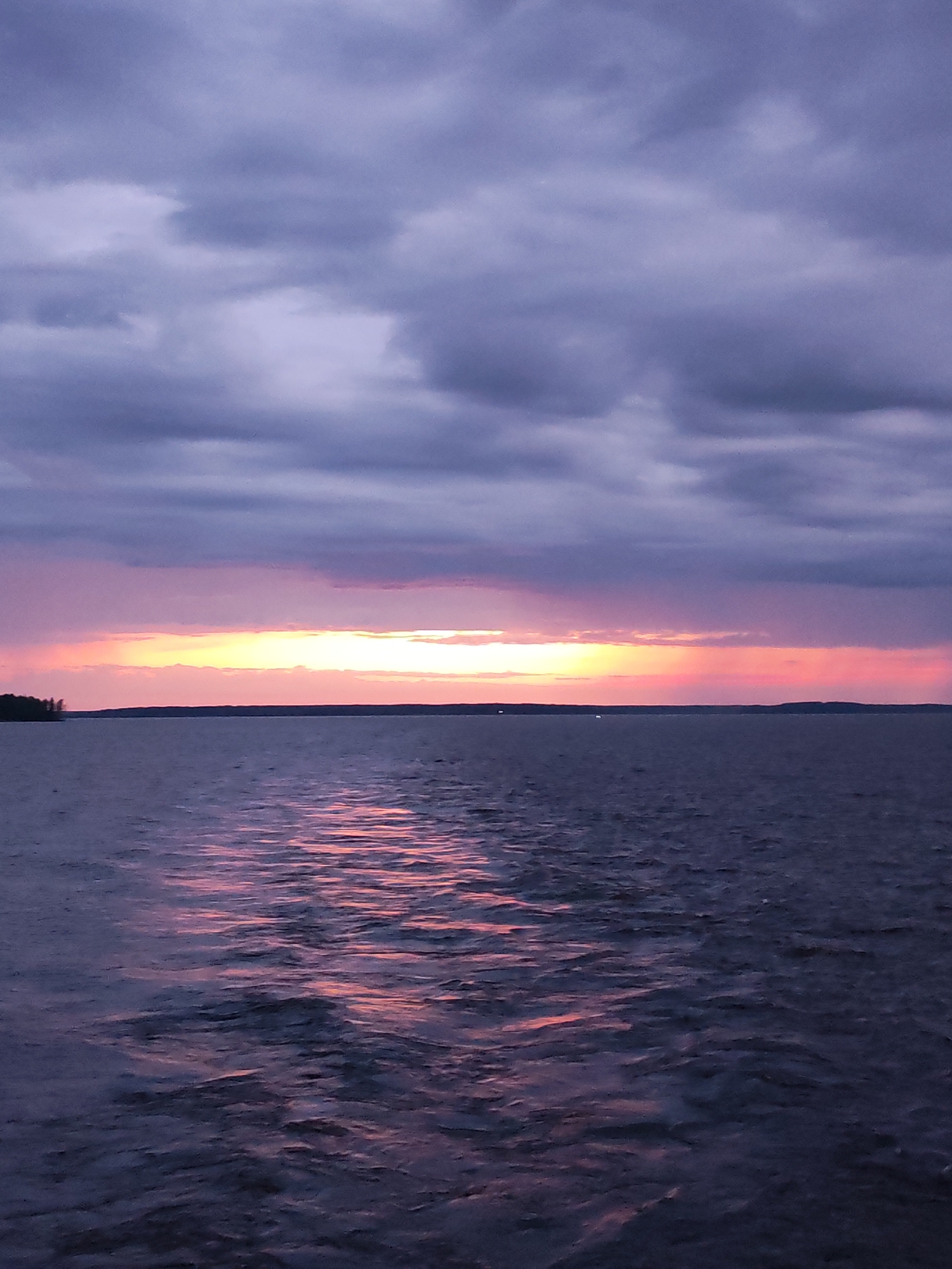 An evening sky over a body of water. The sky is filled with dramatic, layered clouds in shades of deep purple and gray. A band of vibrant sunset colors, including yellow, orange, and pink, is visible on the horizon, breaking through the clouds. The water below is a dark, deep blue with subtle ripples reflecting the colors of the sunset. In the distance, a dark, silhouetted tree line marks the far shore of the water. The overall impression is one of a serene and beautiful natural landscape during twilight. The location is stated to be in the city of Unknown, Russia.