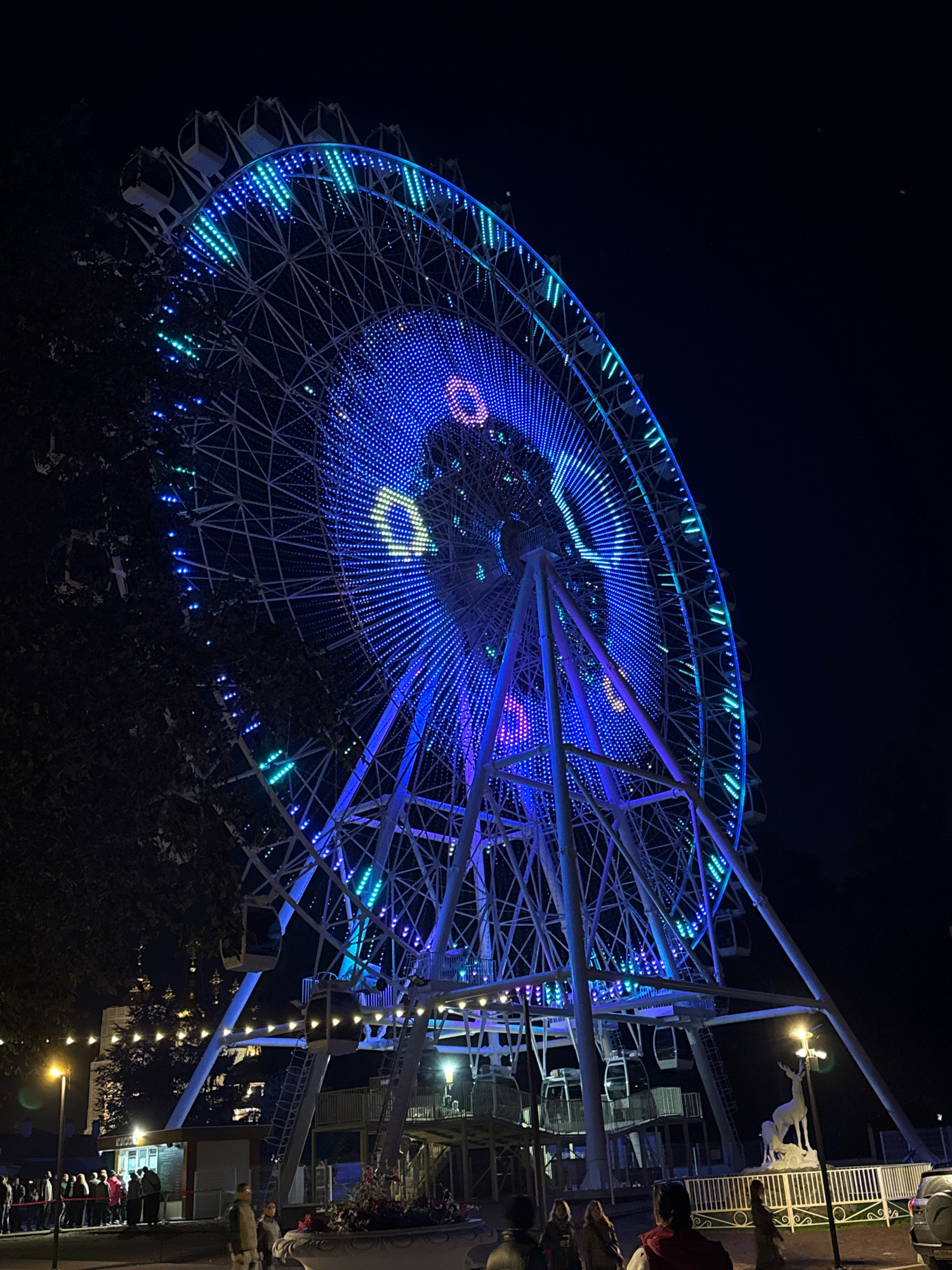 The image depicts a large Ferris wheel illuminated with vibrant blue and green lights at night. The Ferris wheel has several gondolas and a complex metal structure. In the foreground, there are trees on the left side of the image and a crowd of people standing in line near a small building. There are also streetlights and a white statue of a deer and a fawn. In the background, there are more structures and lights, suggesting an amusement park or a similar recreational area. The sky is dark, indicating it is nighttime. The Ferris wheel is the dominant subject, with its lights creating a dazzling display against the dark sky. The scene appears to be a lively public gathering place.